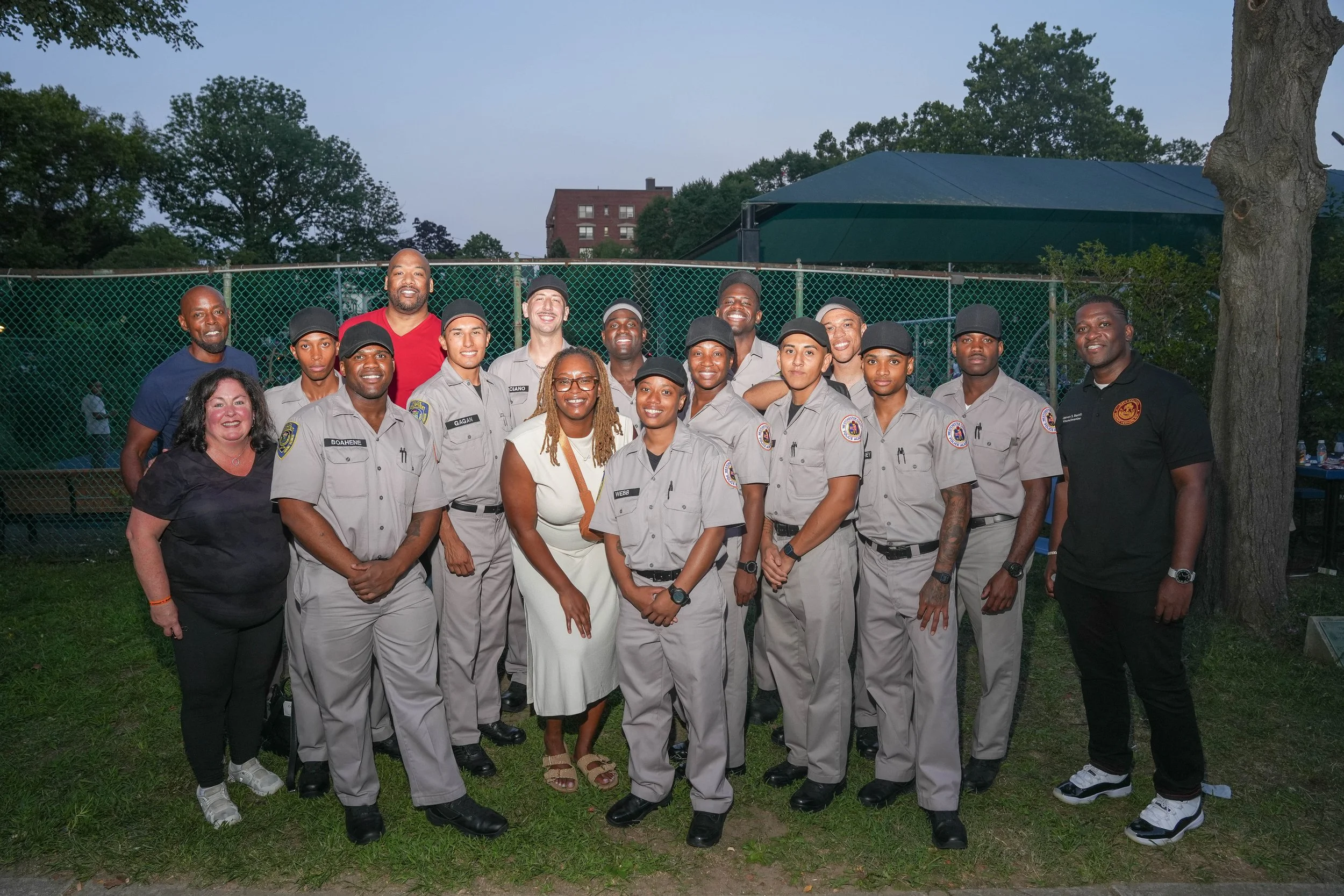 Group of uniformed police officers and civilians posing outdoors in front of a chain-link fence with trees and a building in the background.