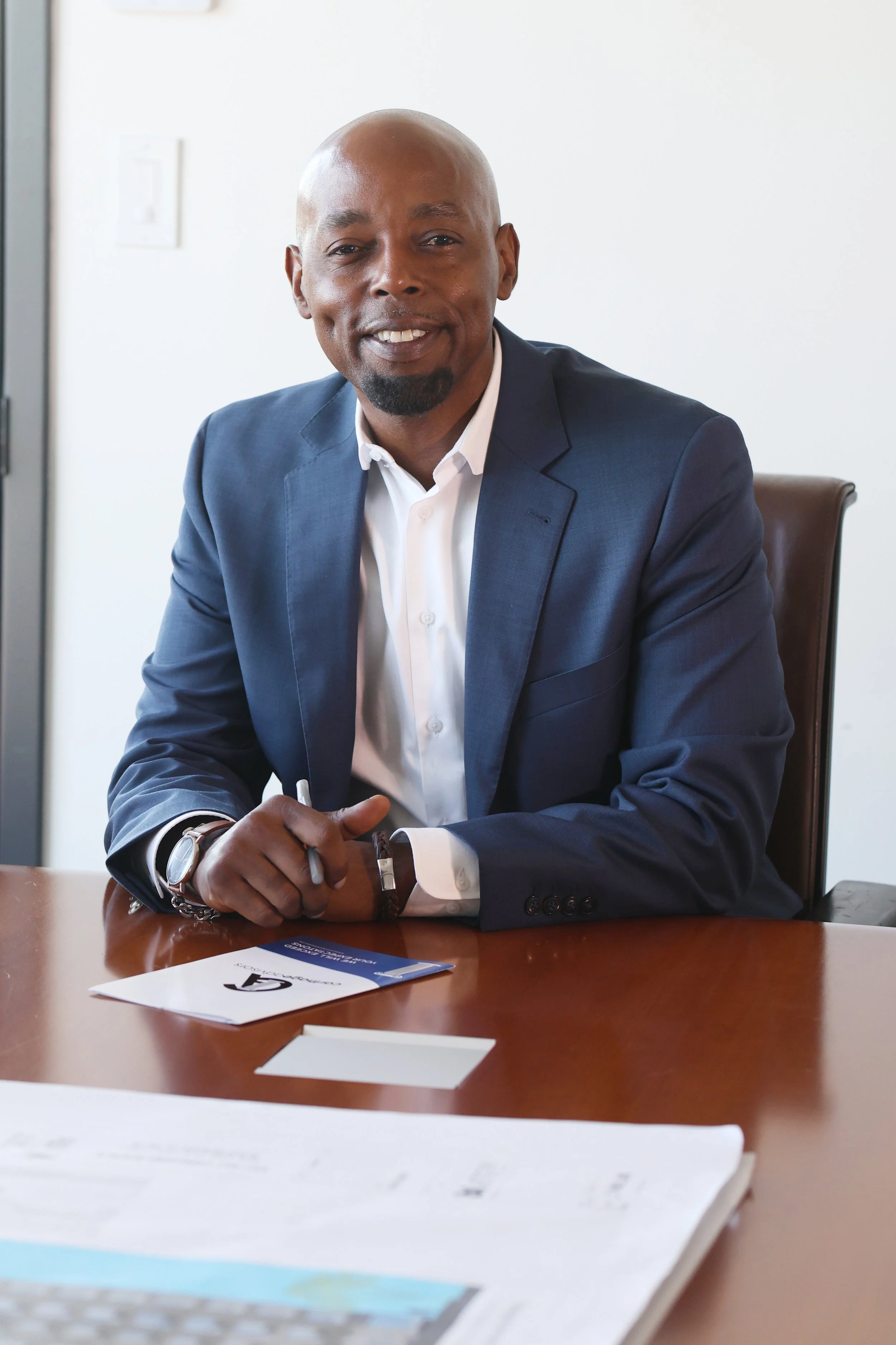 A man in a navy blazer and white shirt, sitting at a desk with business documents, smiling at the camera.