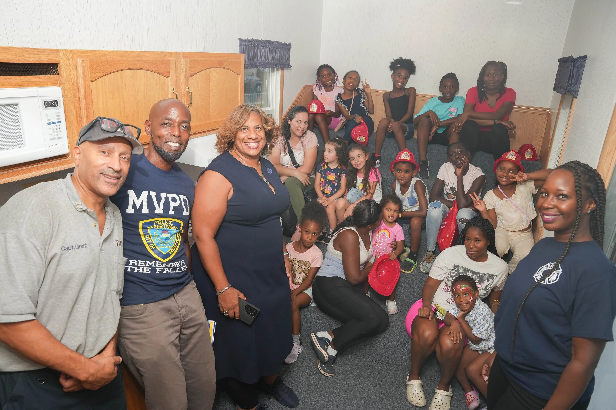 Group of children and adults gathered in a small room, some sitting on the floor and others on a couch, smiling at the camera.