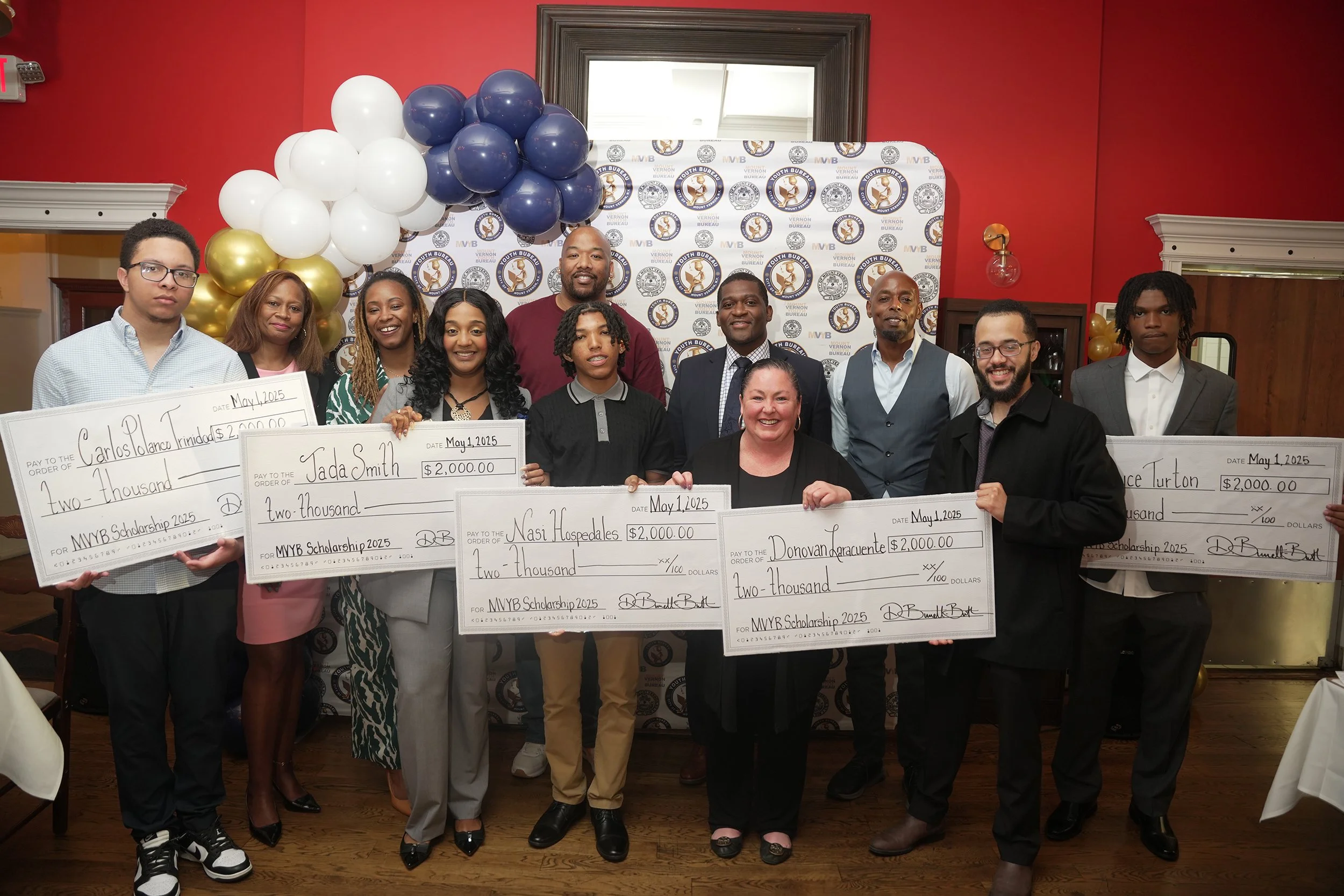 Group of people celebrating at a scholarship award event, holding large ceremonial checks, with balloons and a banner in the background.