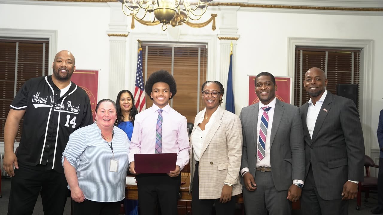 Group of seven diverse people standing together indoors, smiling, with two flags and a chandelier in the background.