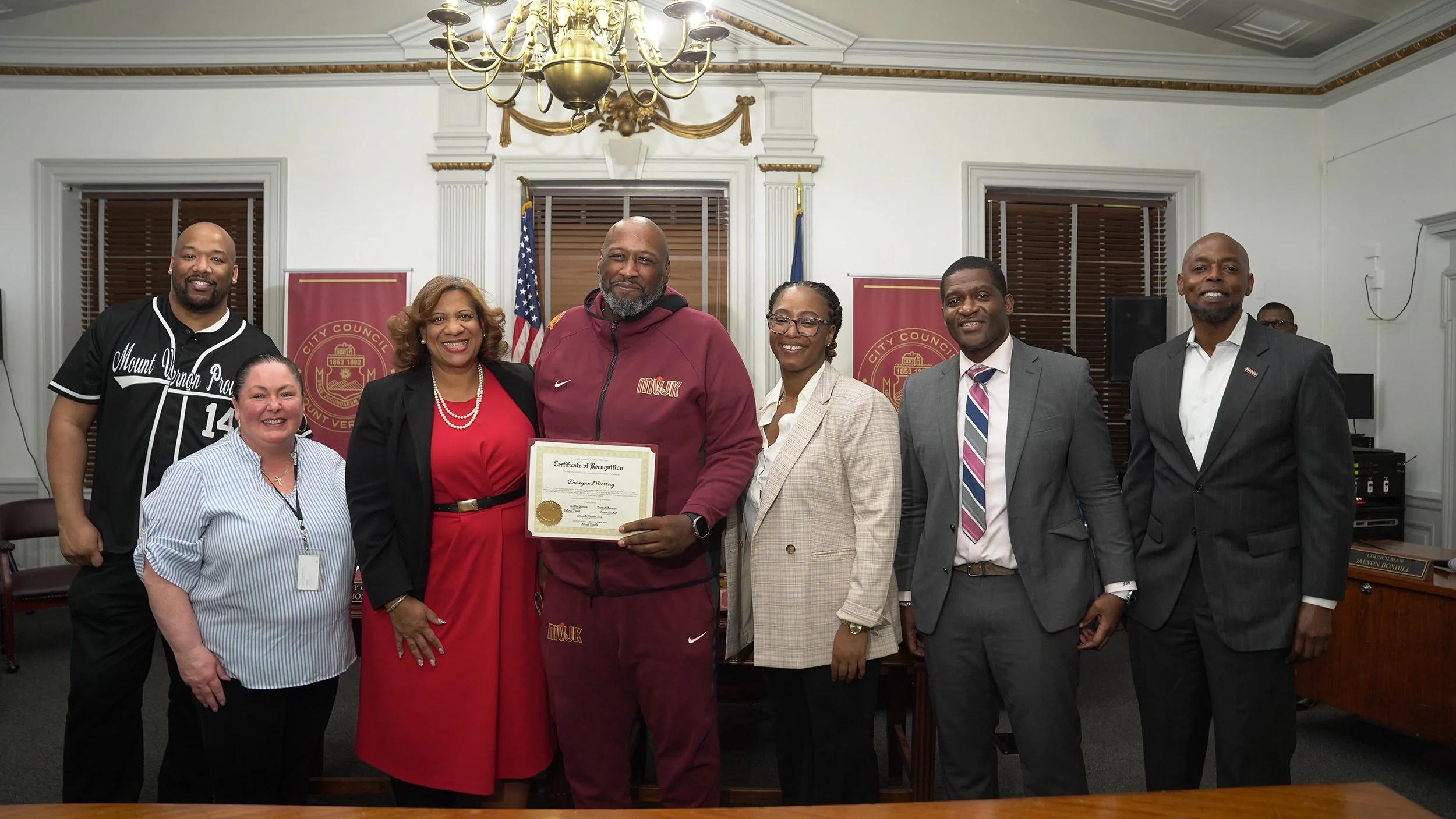 Seven people standing in a room with flags and banners, one holding a framed certificate, celebrating a recognition or achievement.