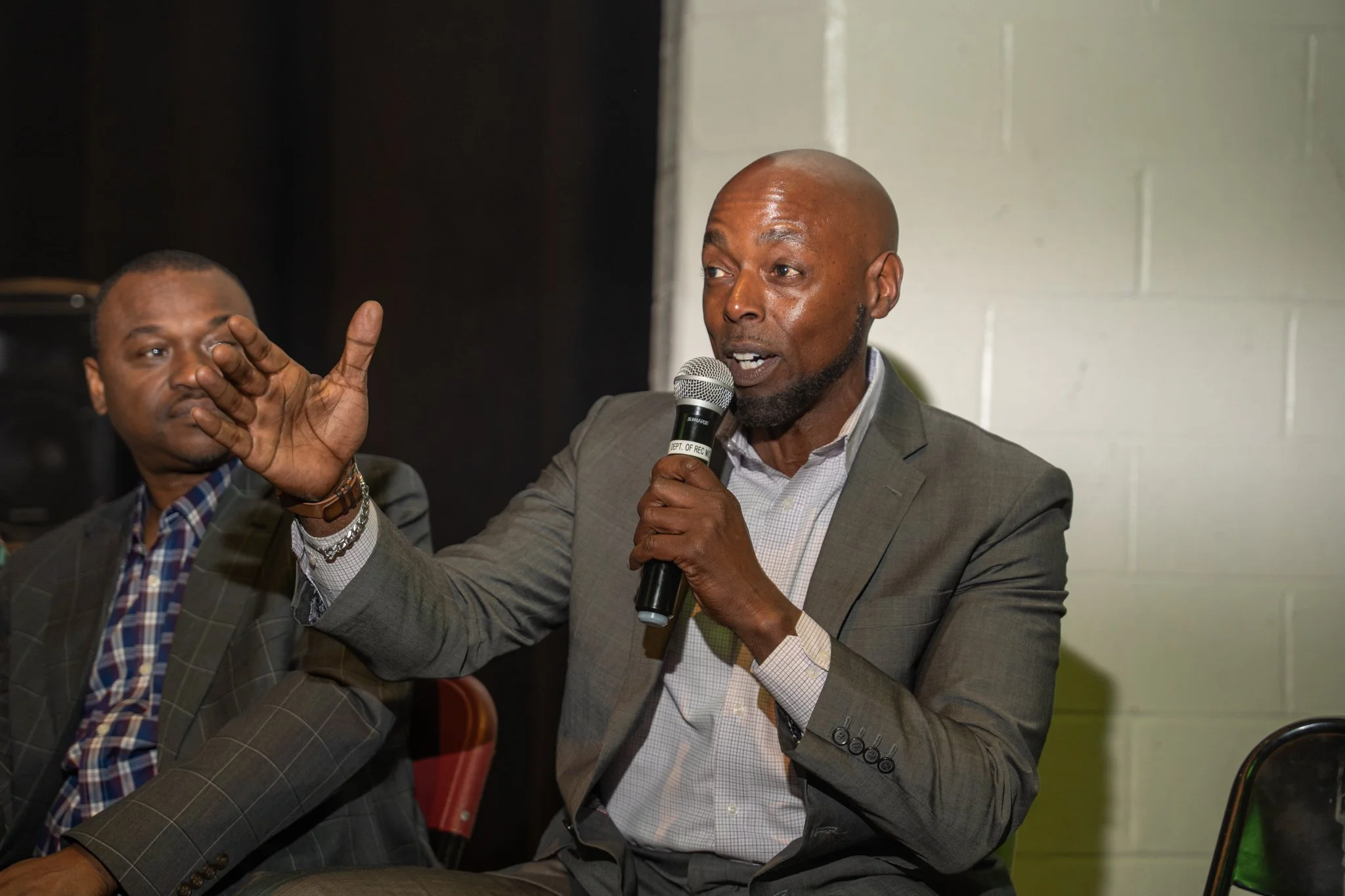 A man in a gray suit and white shirt speaking into a microphone during a discussion or presentation, with another man in a plaid shirt and gray blazer sitting next to him, listening.