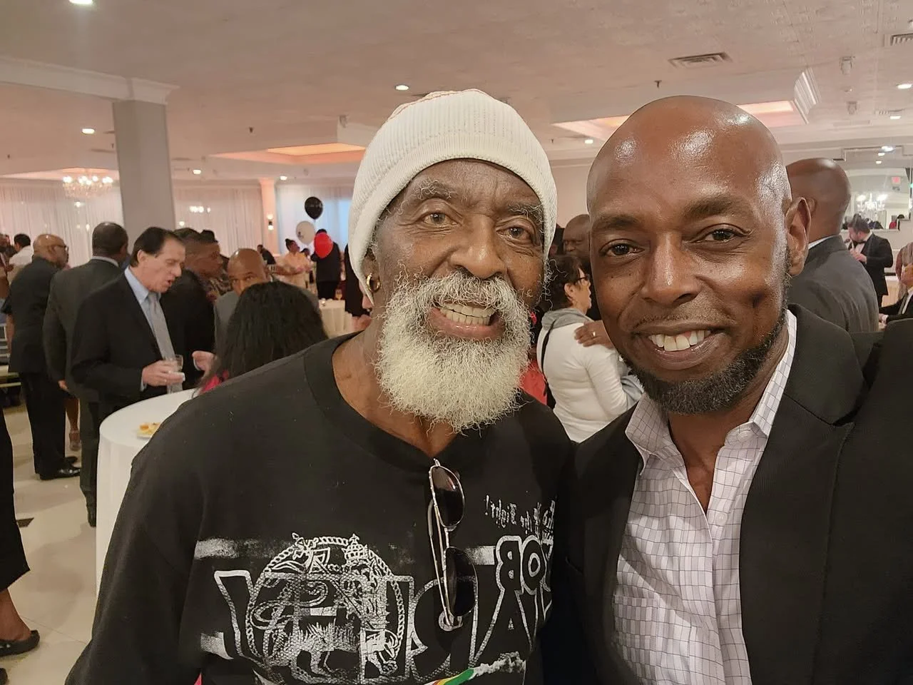 Two smiling men taking a selfie at a social gathering, with other guests mingling in the background.