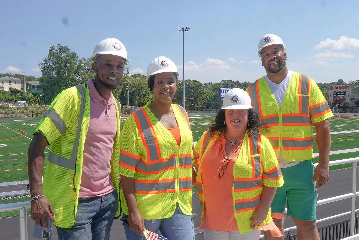 Four construction workers wearing safety vests and helmets stand on a sports field with a track and buildings in the background.