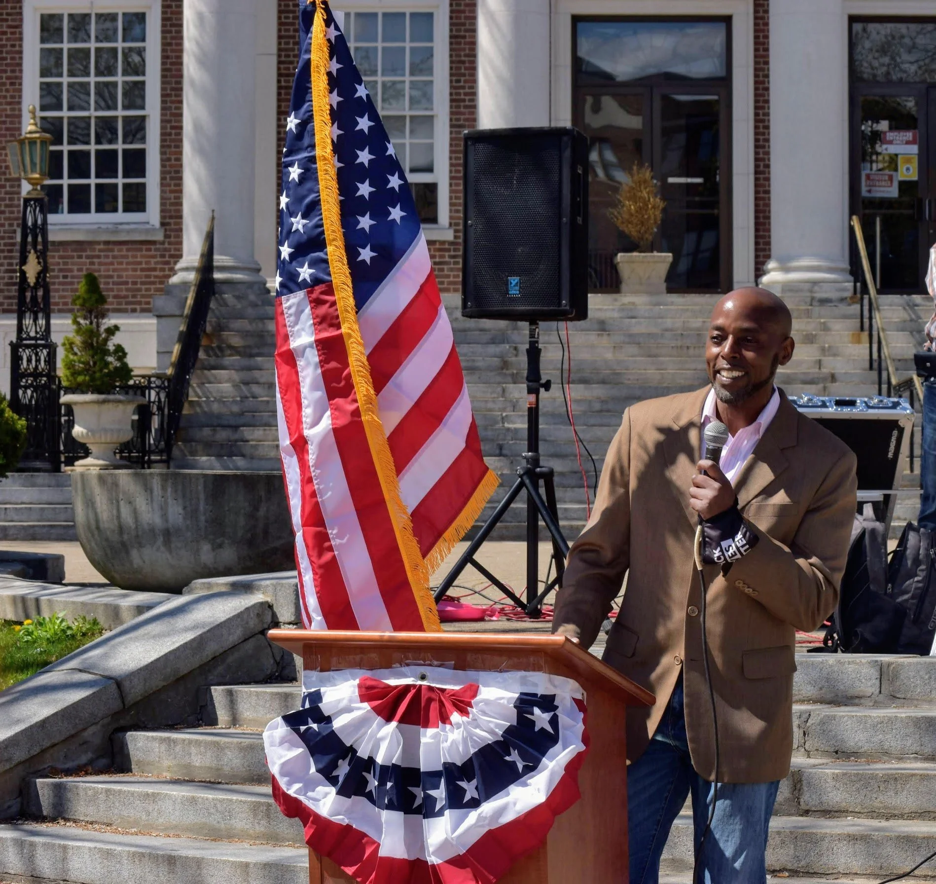 A man in a brown blazer speaking into a microphone at an outdoor event, standing behind a decorated podium with a red, white, and blue bunting. An American flag is nearby, and there are steps leading up to a building with columns and large windows in