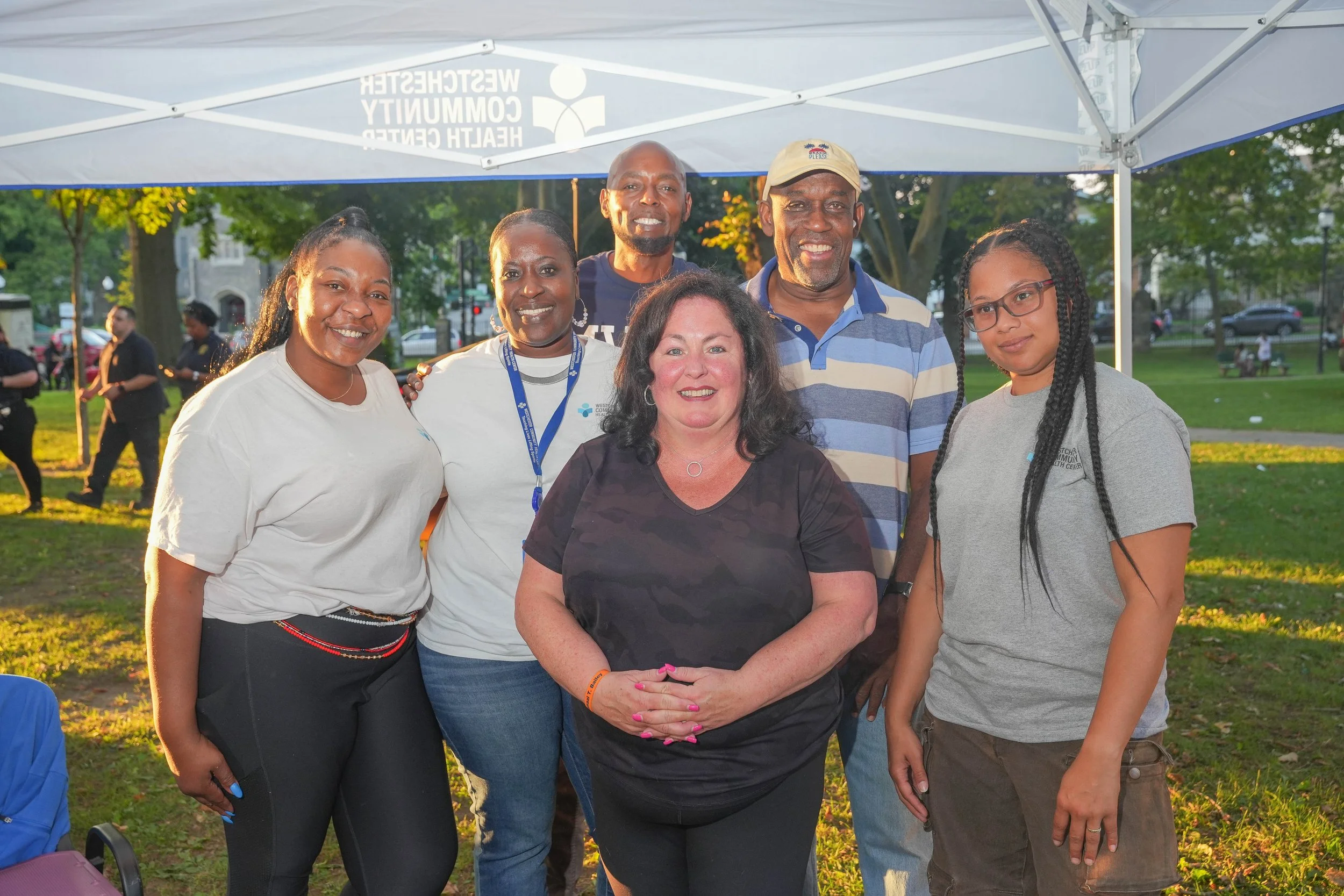 Group of seven diverse people outdoors under a white canopy, smiling at the camera, with trees and park setting in the background.