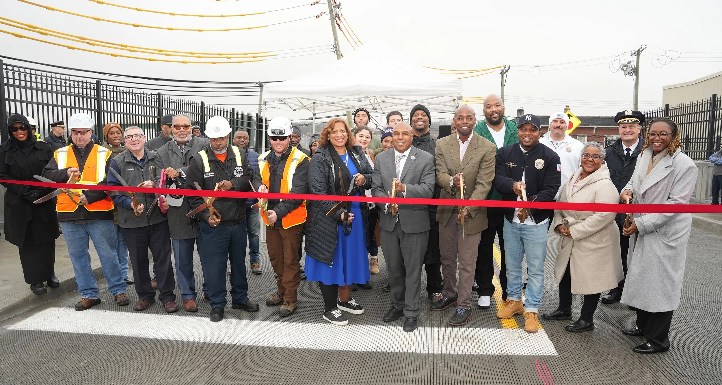 A diverse group of people at a ribbon-cutting ceremony, holding scissors and standing together outdoors on a road, with some wearing safety vests, hard hats, or formal attire.