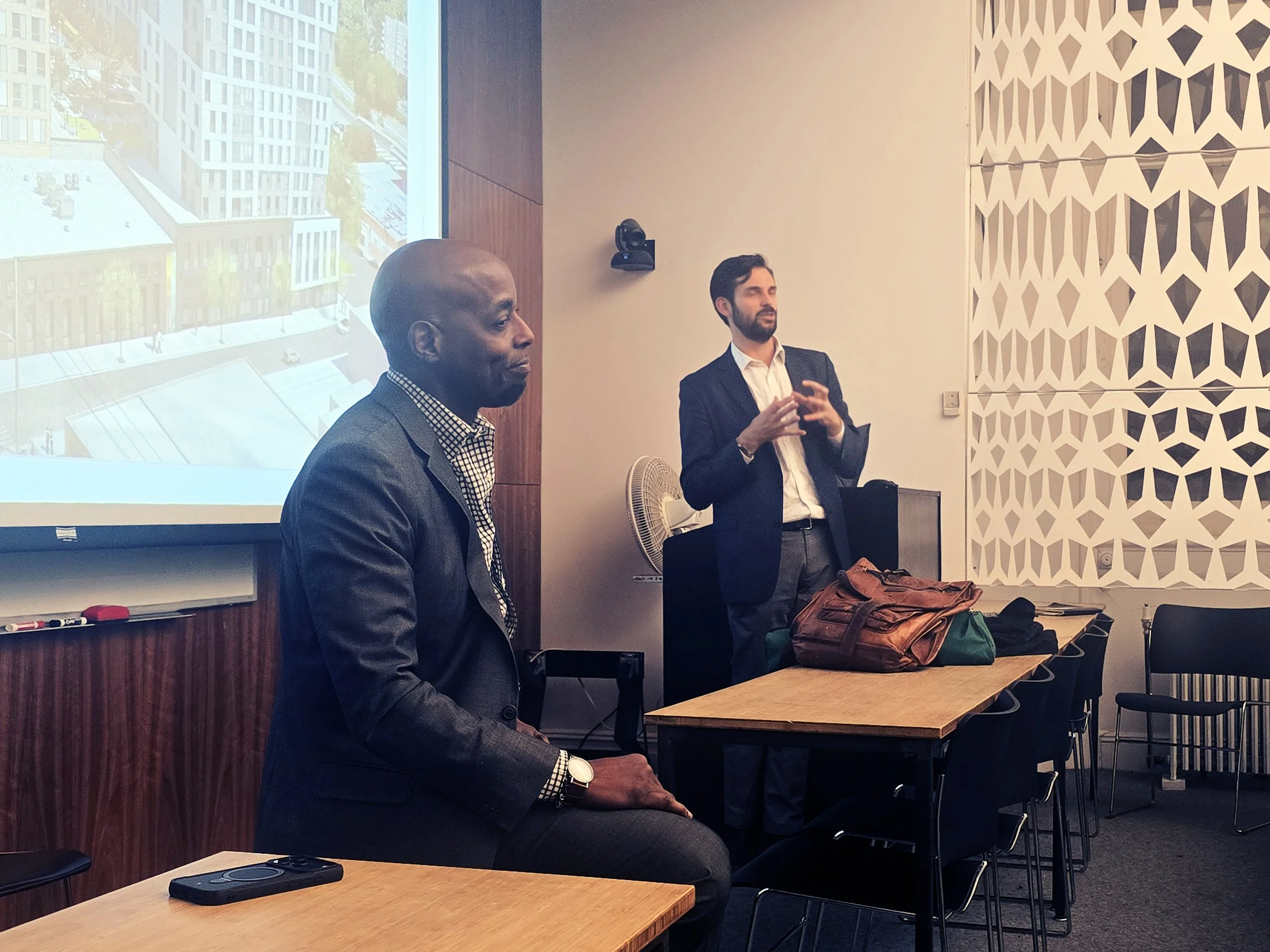 Two men in suits speaking in a conference room, one standing and the other sitting, with a projector screen showing a building exterior behind them.