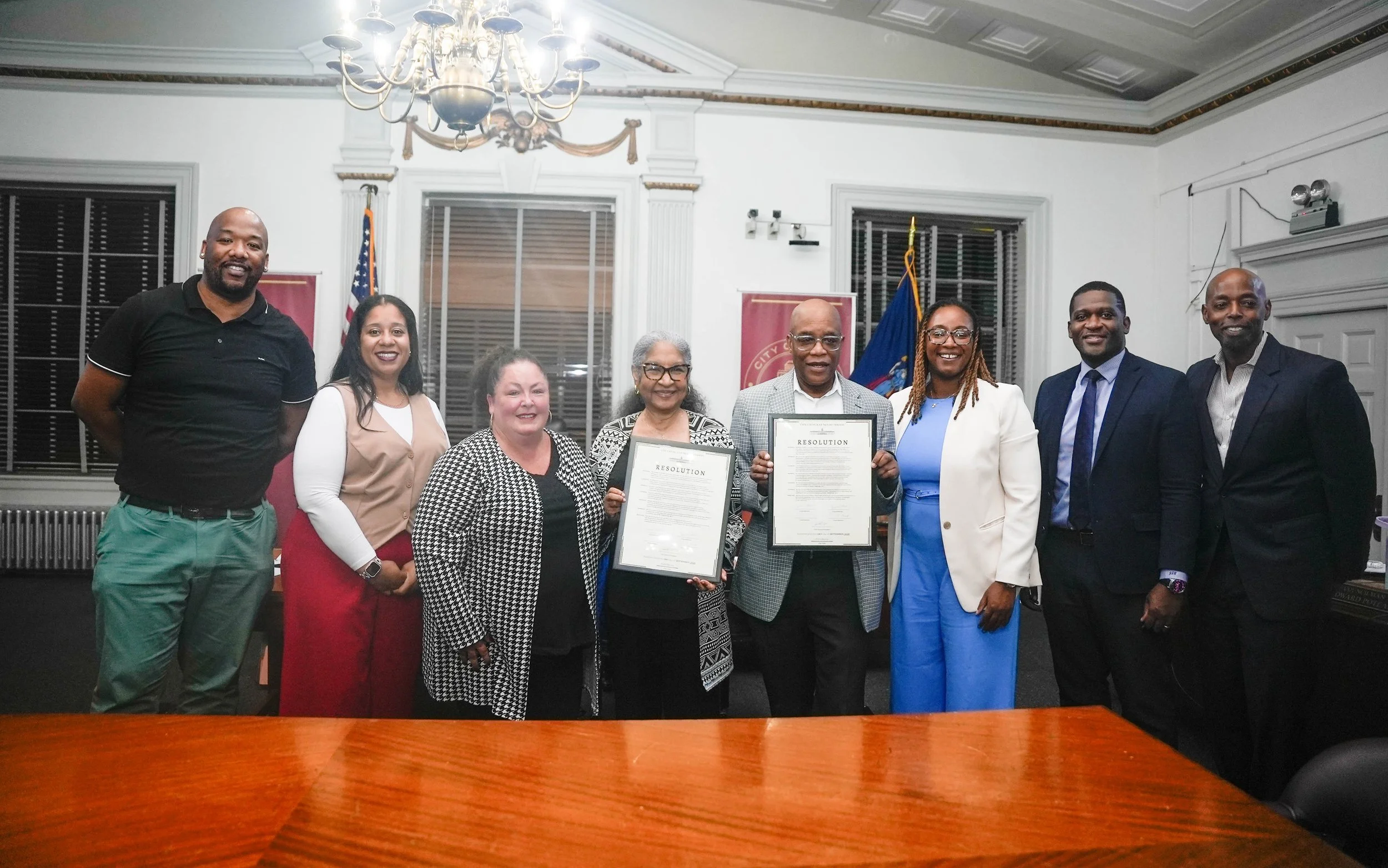 Group of nine people standing in a room with official flags and framed documents, some holding signed resolutions, with a wooden table in the foreground and a chandelier overhead.