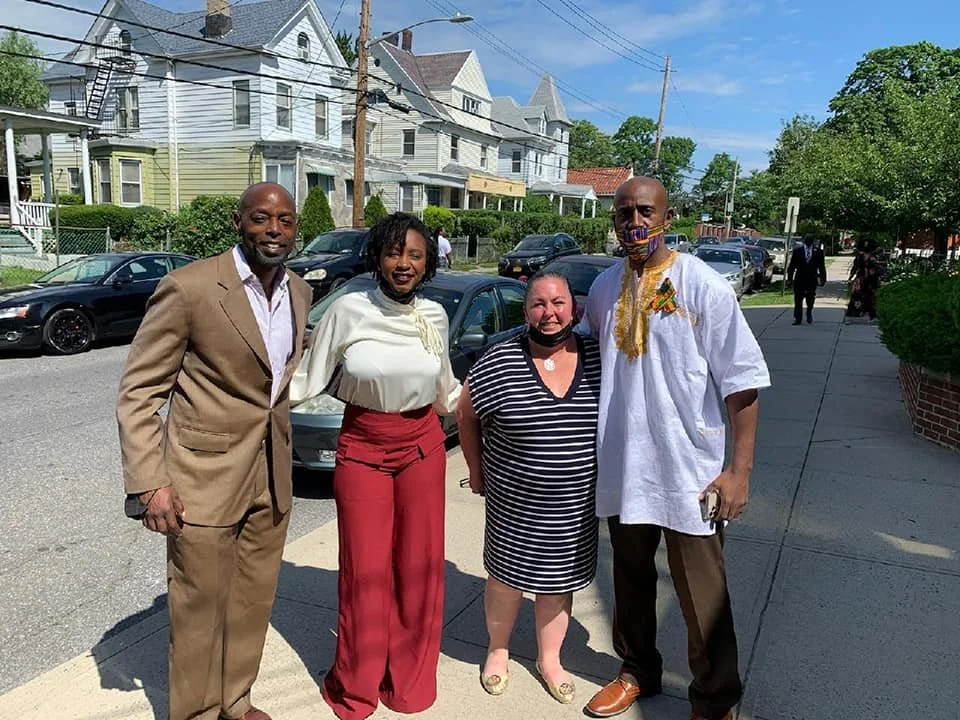 Four people standing on a sidewalk in a neighborhood, smiling at the camera, with houses and cars in the background.