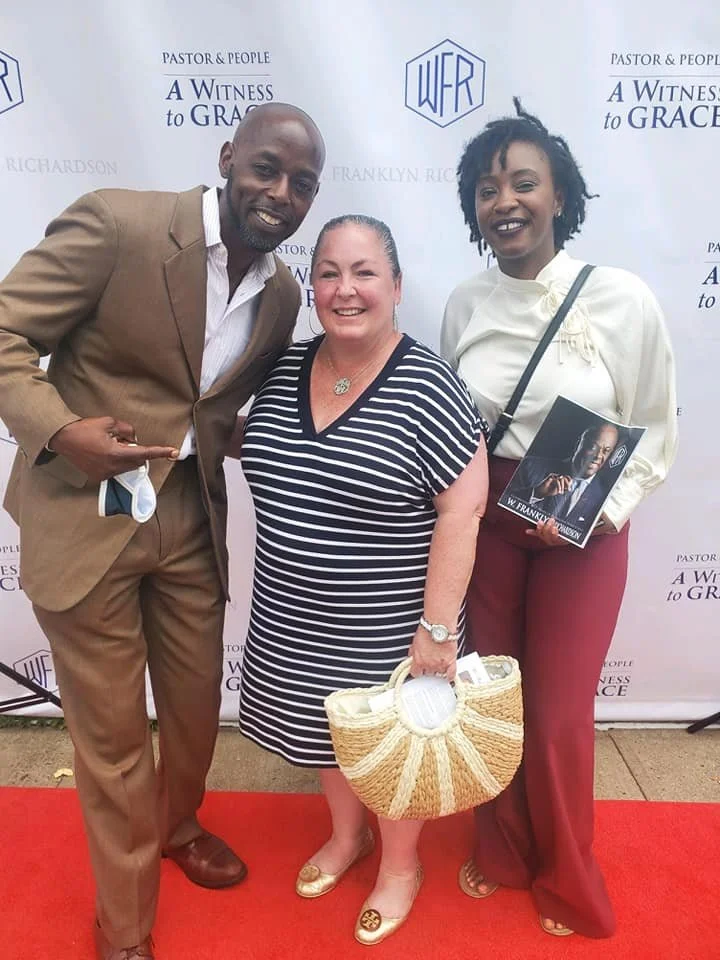 Three people, two women and one man, standing on a red carpet in front of a backdrop with logos and text. The woman in the middle is holding a woven bag and smiling, the man on the left is pointing at her and smiling, and the woman on the right is ho