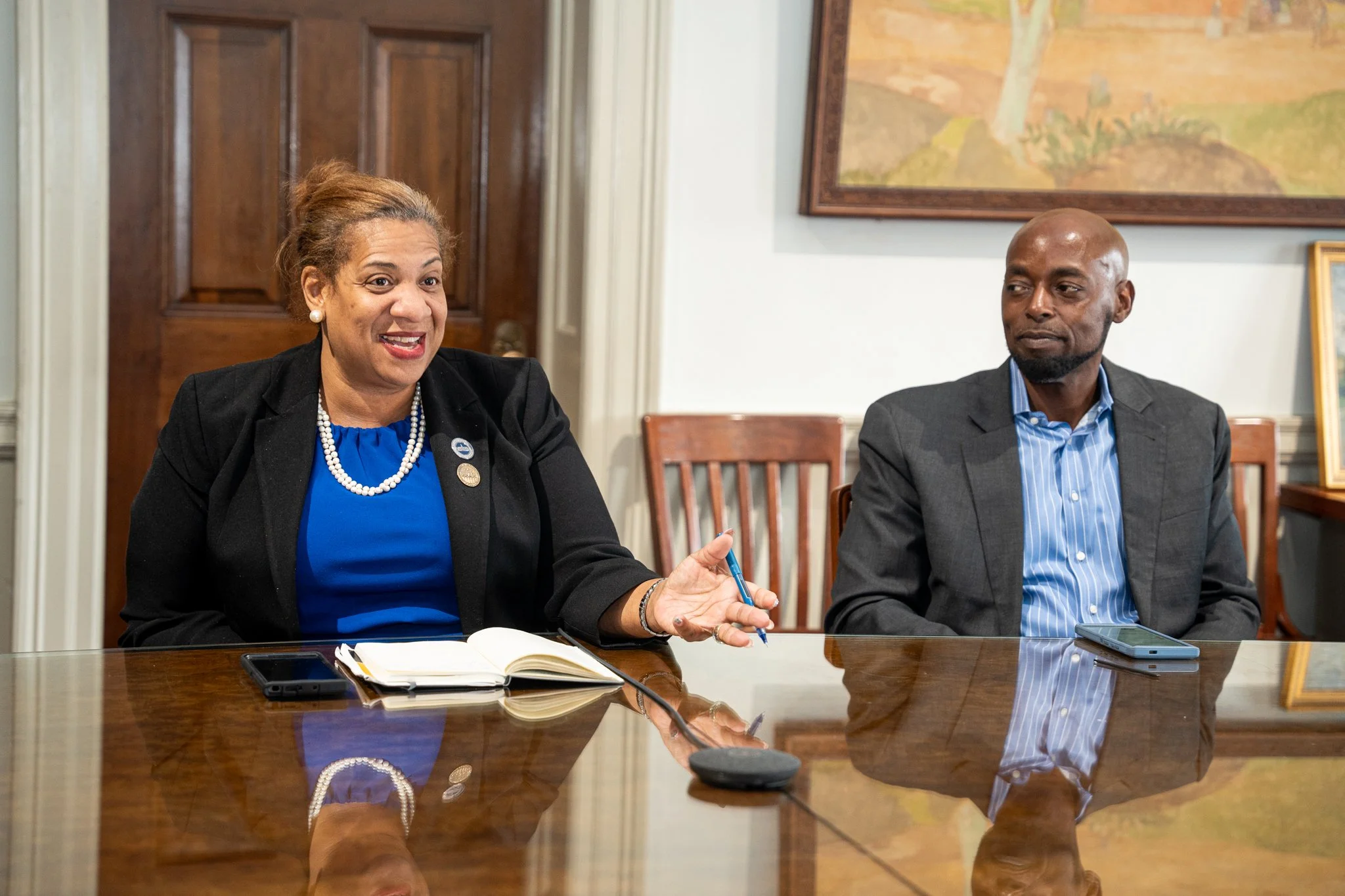 Two people, a woman and a man, sitting at a conference table in a formal setting, engaged in a conversation, with documents and a smartphone on the table.