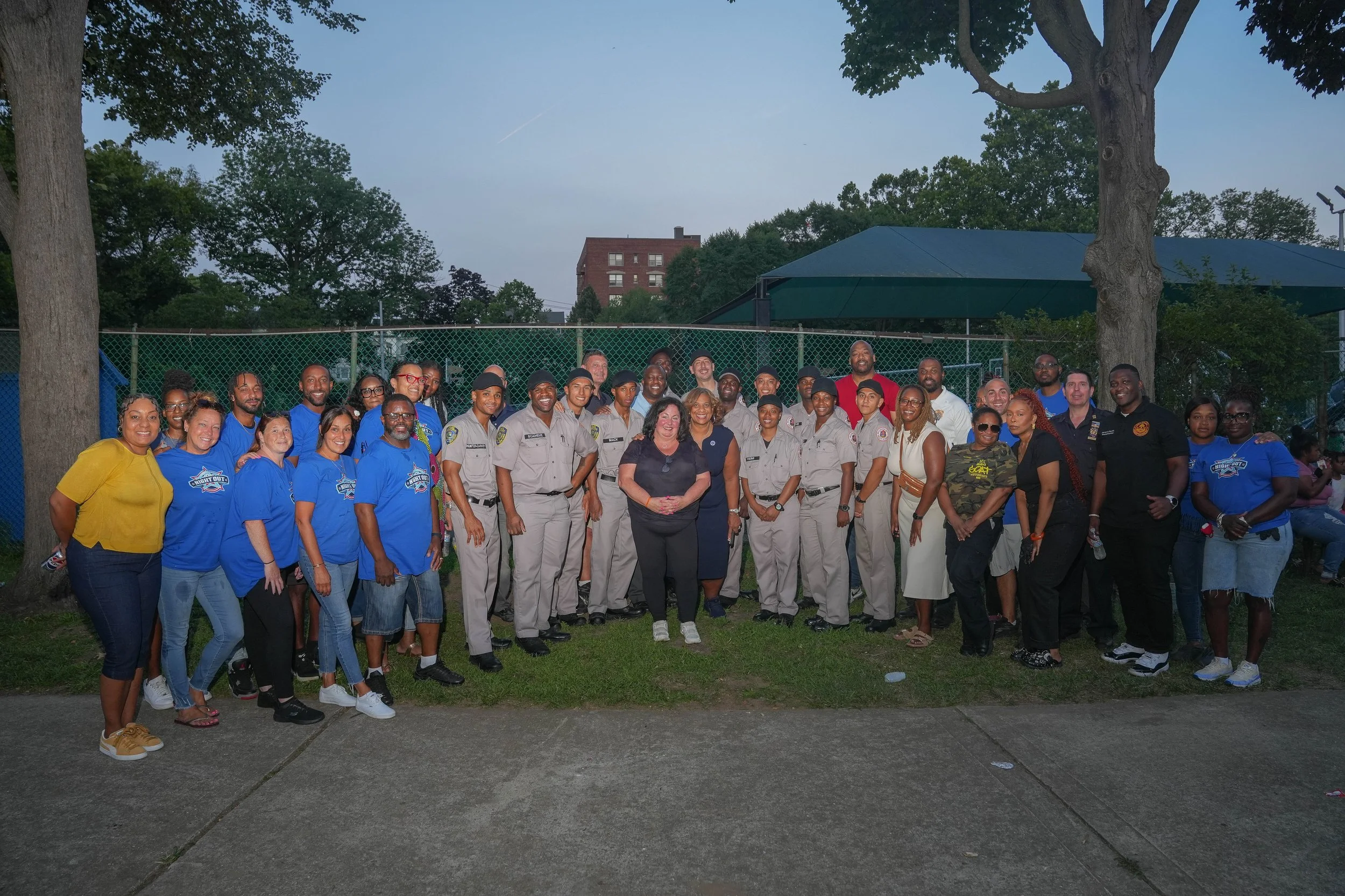 A large group of people posing outdoors near trees and a chain-link fence, some in police uniforms and others in casual clothing, during the evening.