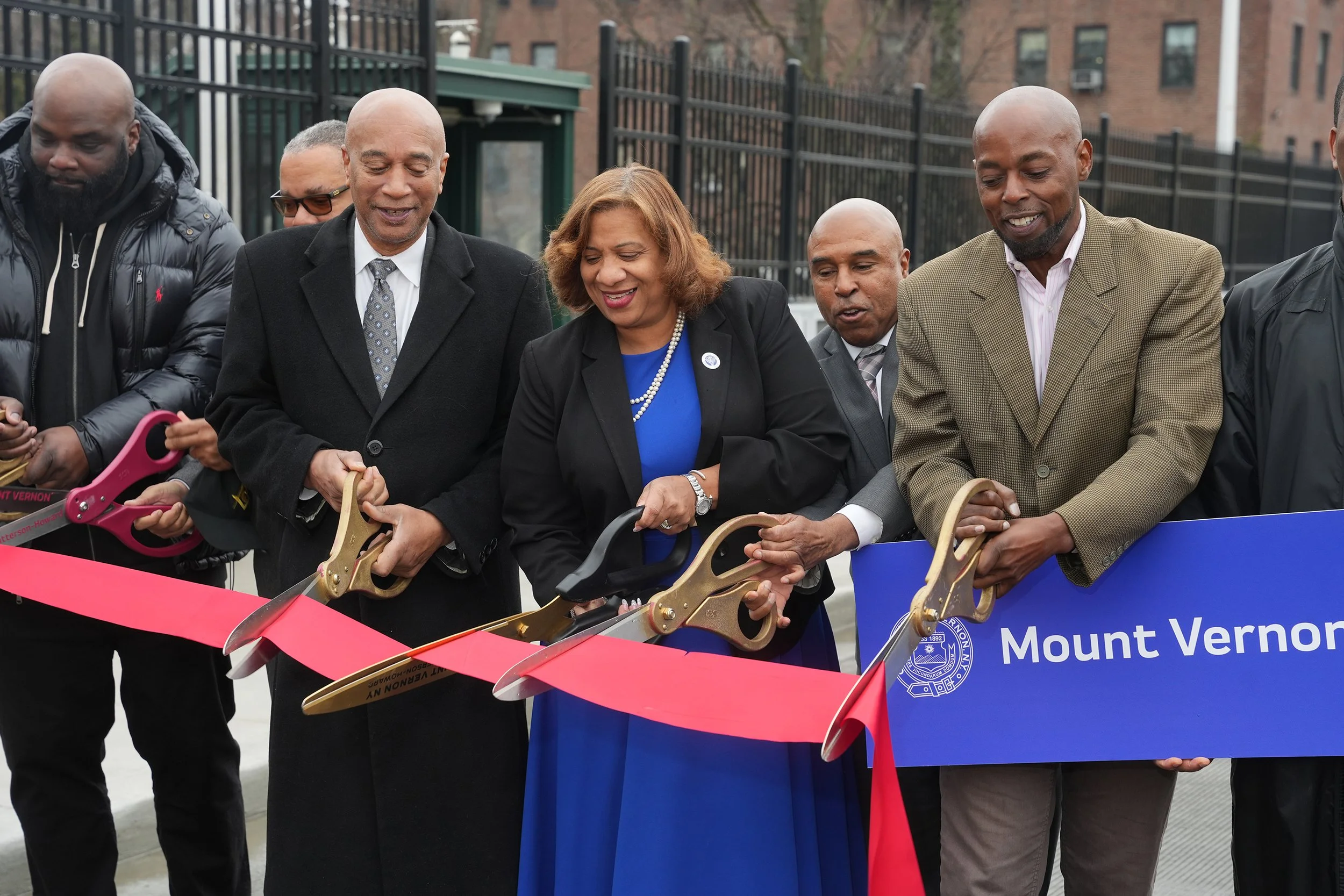 Group of people at a ribbon-cutting ceremony holding scissors to cut a red ribbon, with a blue sign that says 'Mount Vernon'.