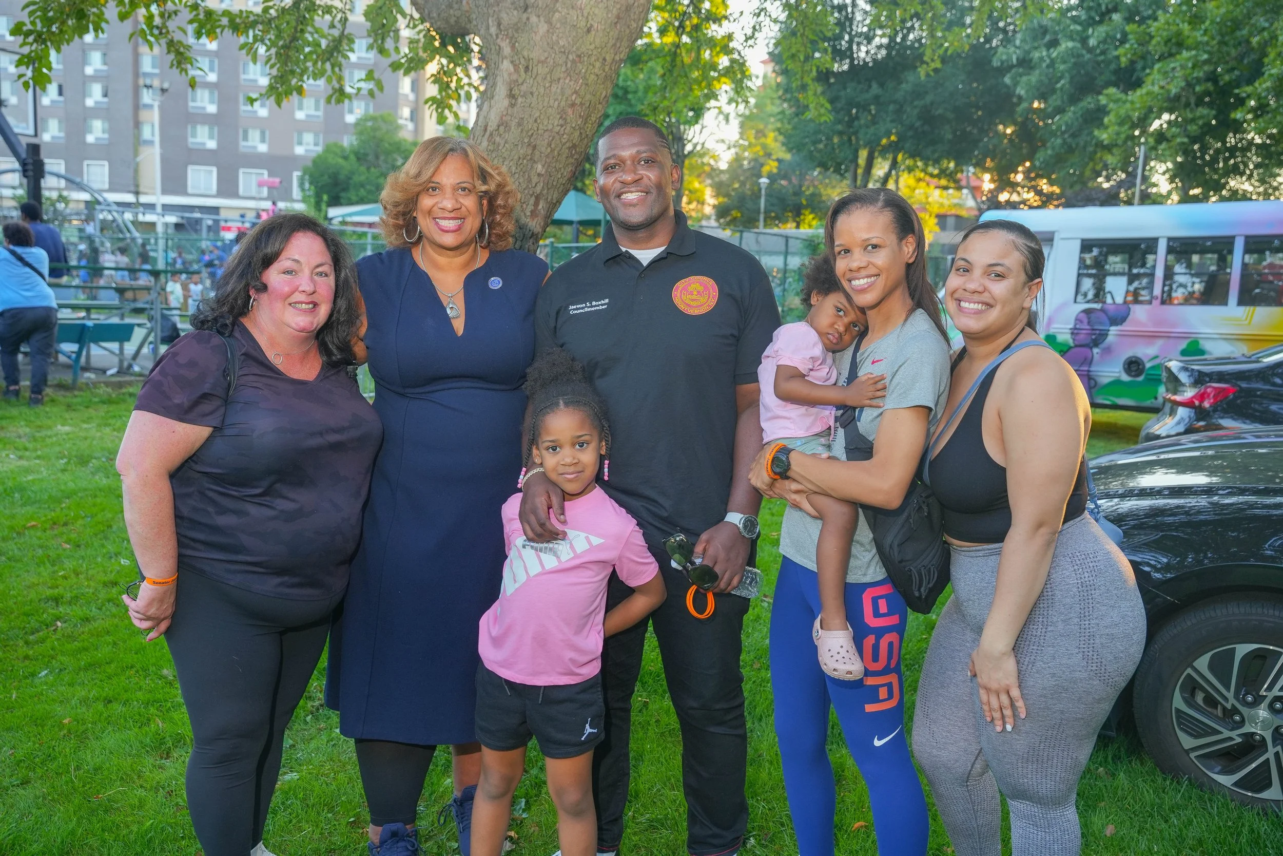 Group of six women and two young girls standing outdoors at a park, smiling. One woman holds a small girl in her arms, while a young girl stands in front of a man, and another girl stands between two women.