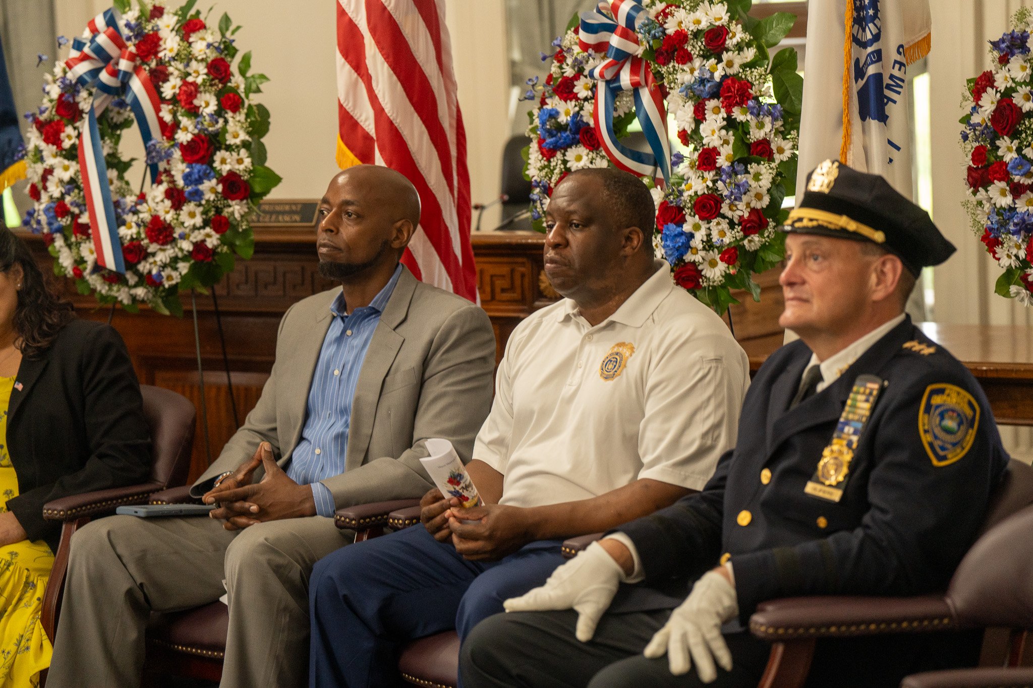 Three men sitting in a formal setting with American flags and floral arrangements in the background. The man on the right is a police officer in uniform, the man in the middle wears a white polo shirt with a badge, and the man on the left is in a lig