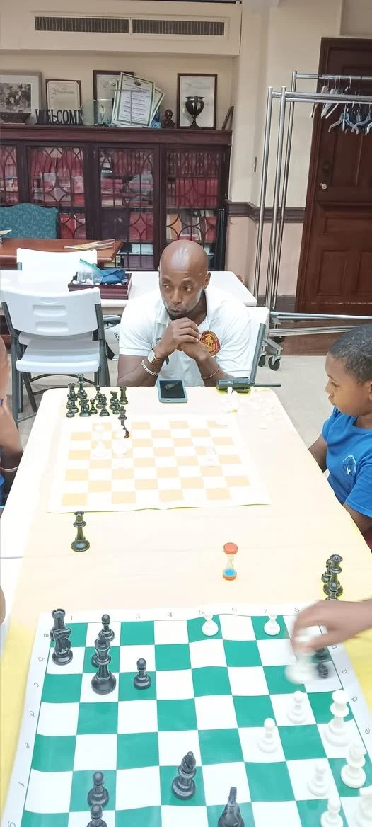 A man and a boy playing chess at a table in a room with shelves and framed certificates, with a chessboard and a separate game piece on the table.
