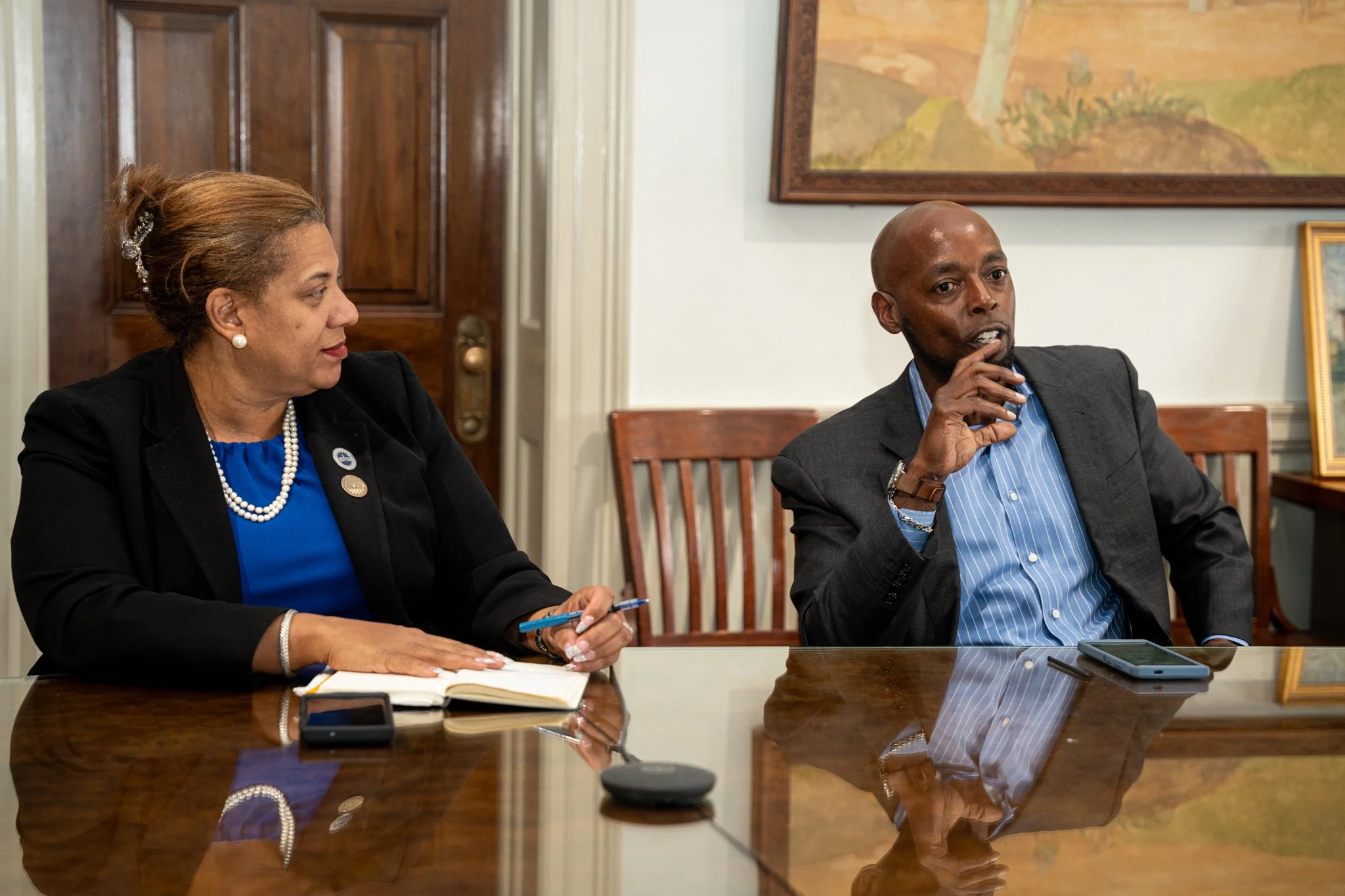 A woman and a man sitting at a conference table during a meeting or discussion. The woman is wearing a blue top, black blazer, pearl necklace and earrings, and holding a pen, with a notebook and phone on the table. The man is wearing a dark blazer, l