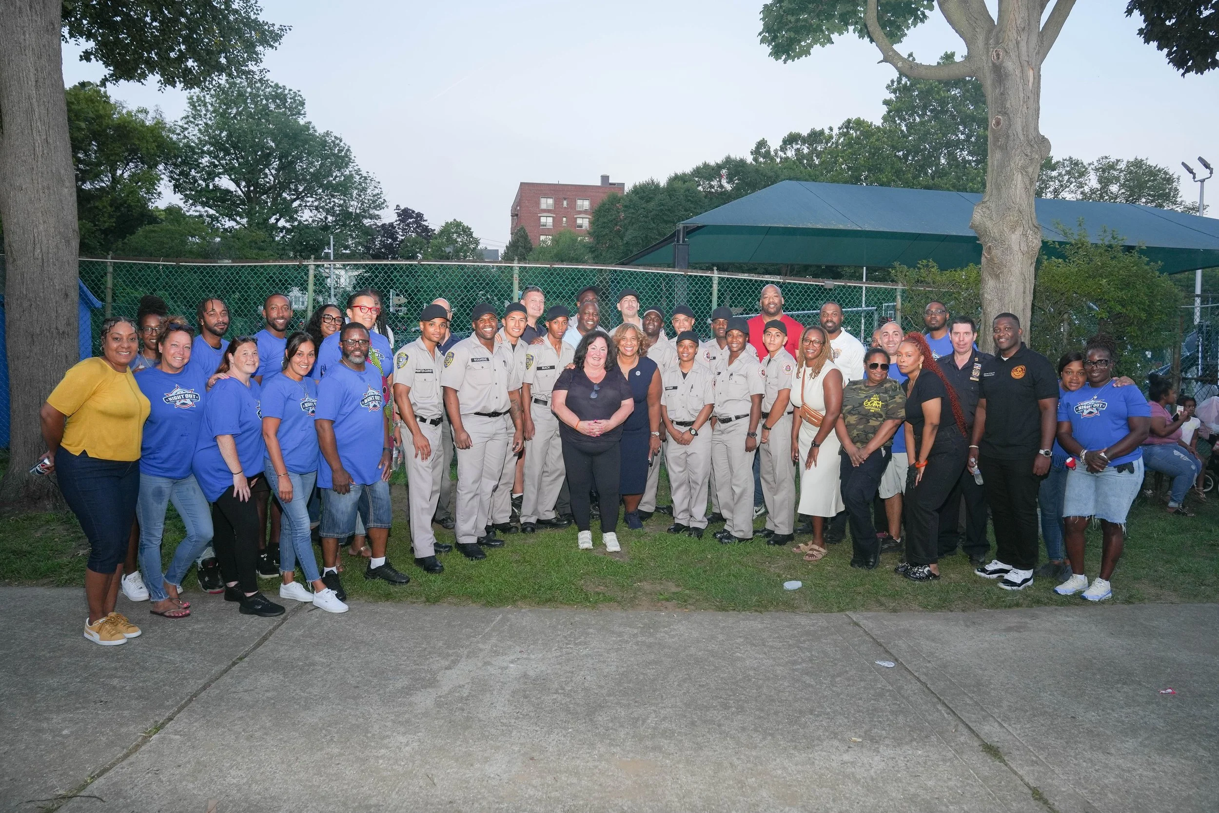A large group of people, including police officers, community members, and possibly officials, gathered outdoors in a park-like setting for a group photo. Some are wearing police uniforms, others in casual or community organization clothing, standing
