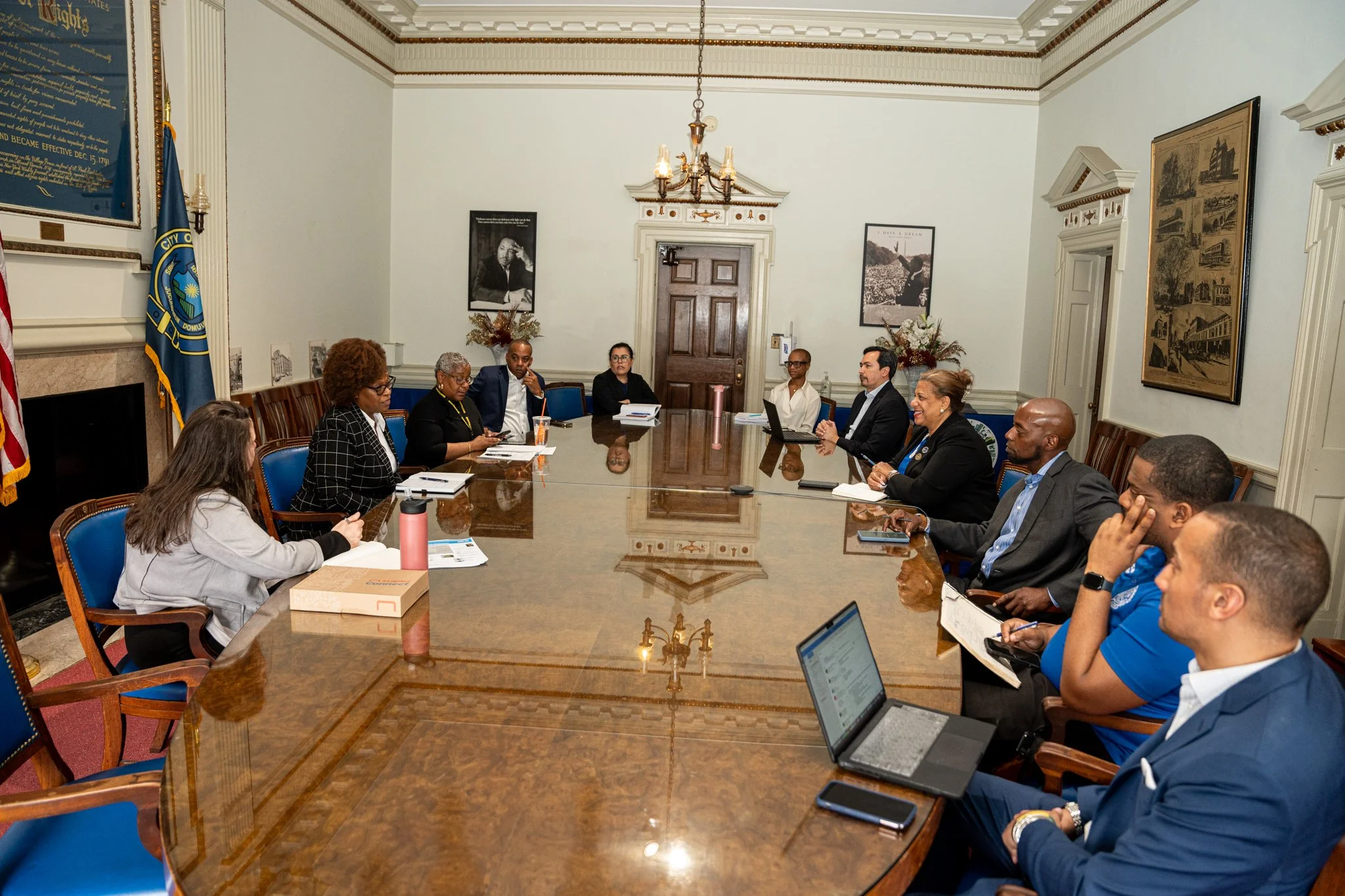 A diverse group of people sit around a large, polished wooden conference table in a formal meeting room with framed photographs on the walls and flags in the corner.