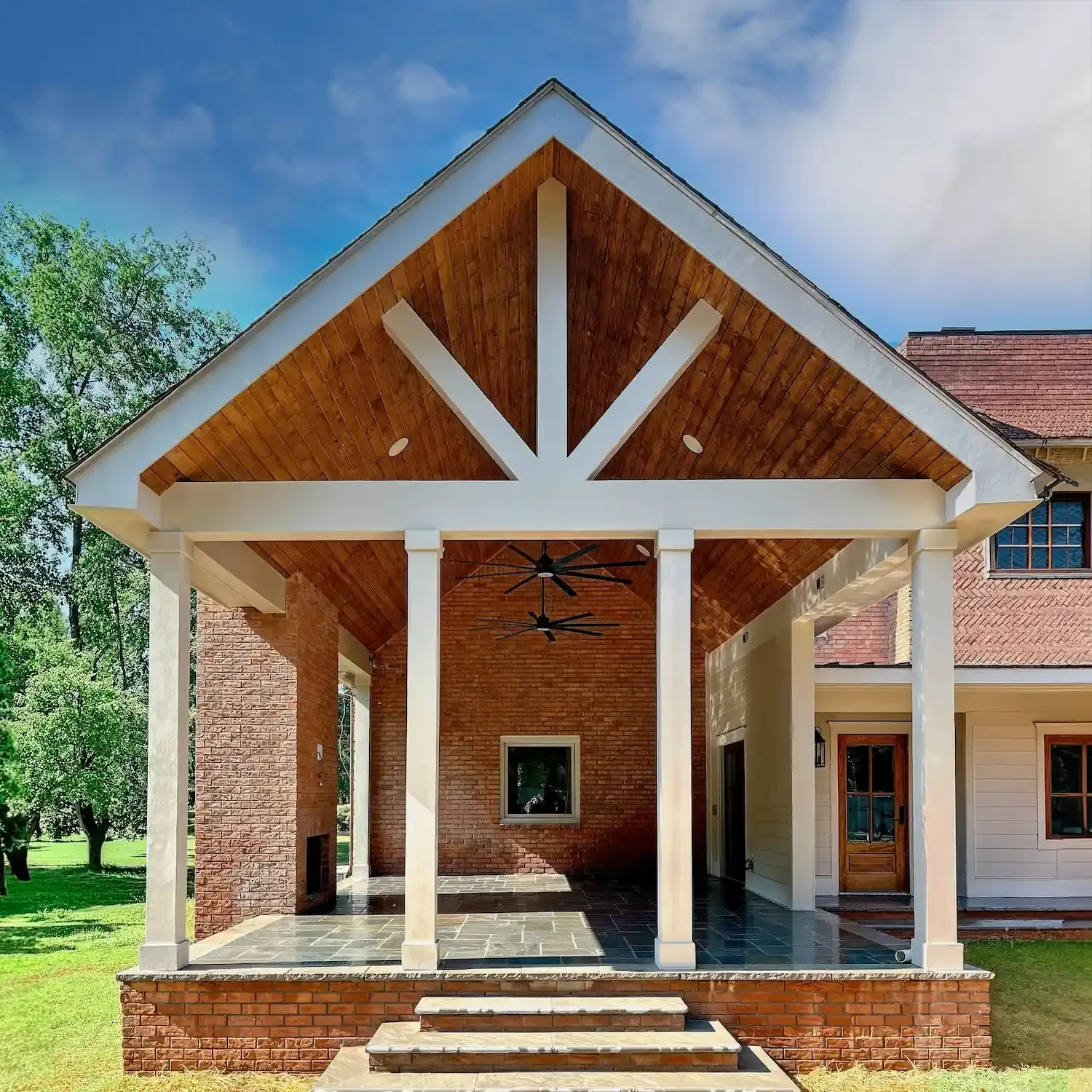 Modern covered patio with tall white columns, wood gabled ceiling, large black ceiling fans, and dark stone tile steps on a red brick home exterior in Hendersonville, Tennessee.