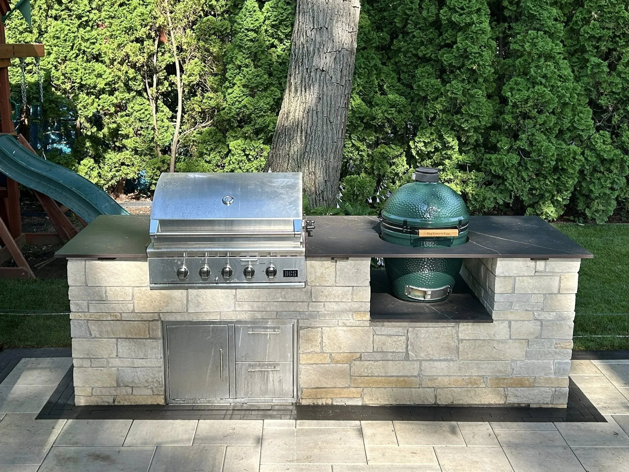An outdoor kitchen area with a built-in stainless steel grill, a green Weber kettle grill, and a stone counter, surrounded by green trees and a grass lawn.
