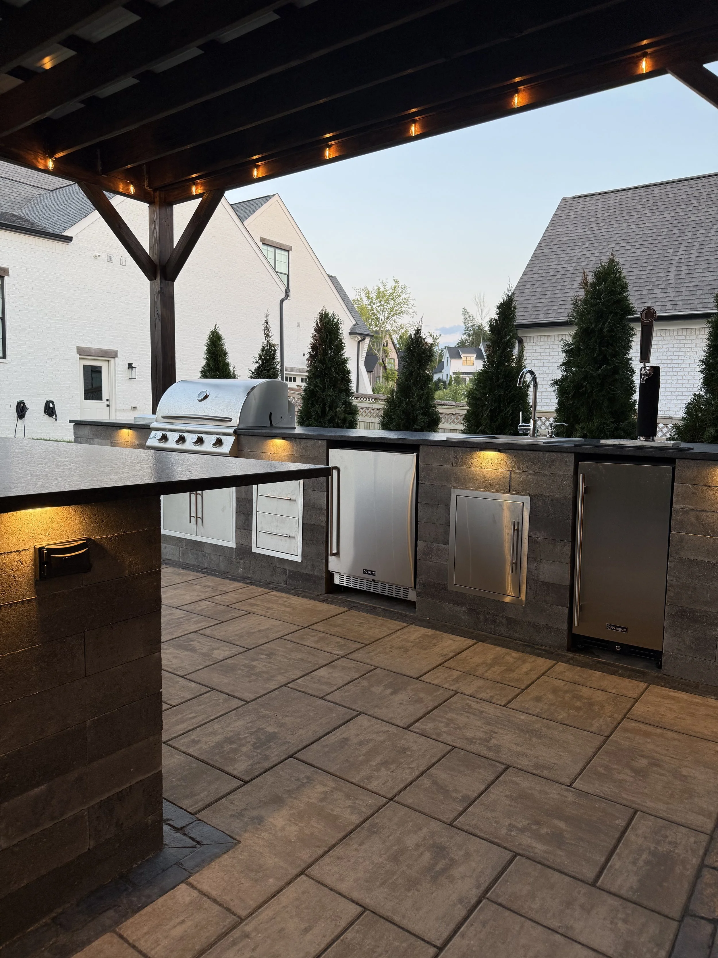 Outdoor kitchen area with a built-in grill, stainless steel cabinets, a sink, and a tile countertop under a wooden pergola with string lights, overlooking neighboring houses and trees.