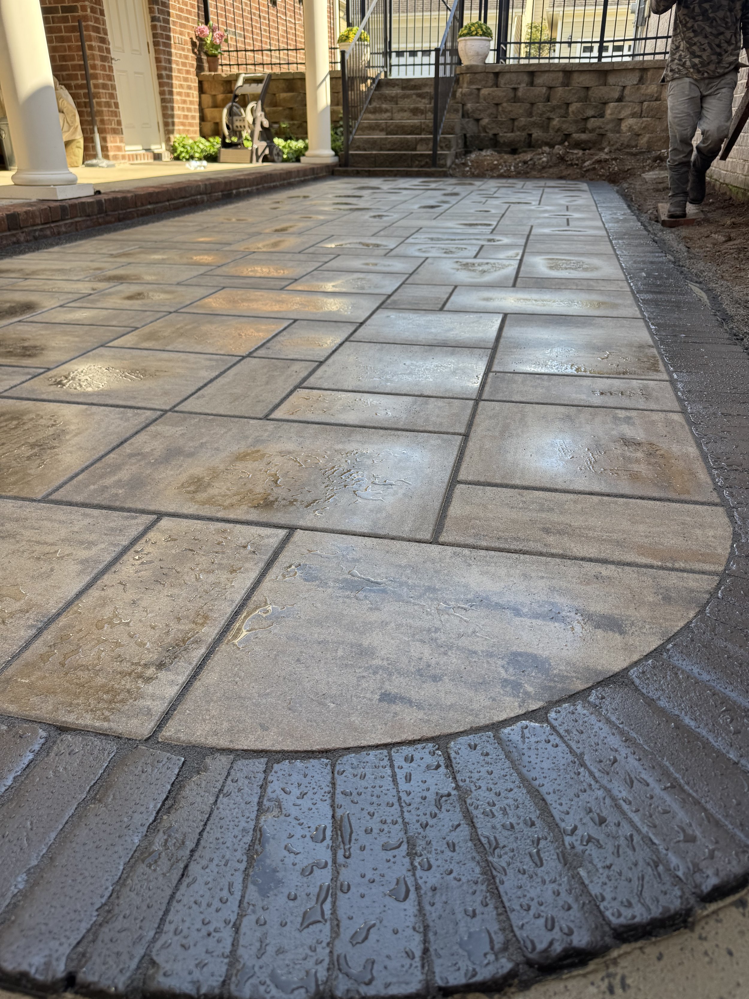 Newly paved concrete driveway with wet surface, bordered by brick edges and a darker brick border, with construction in progress on the right side and a person working.