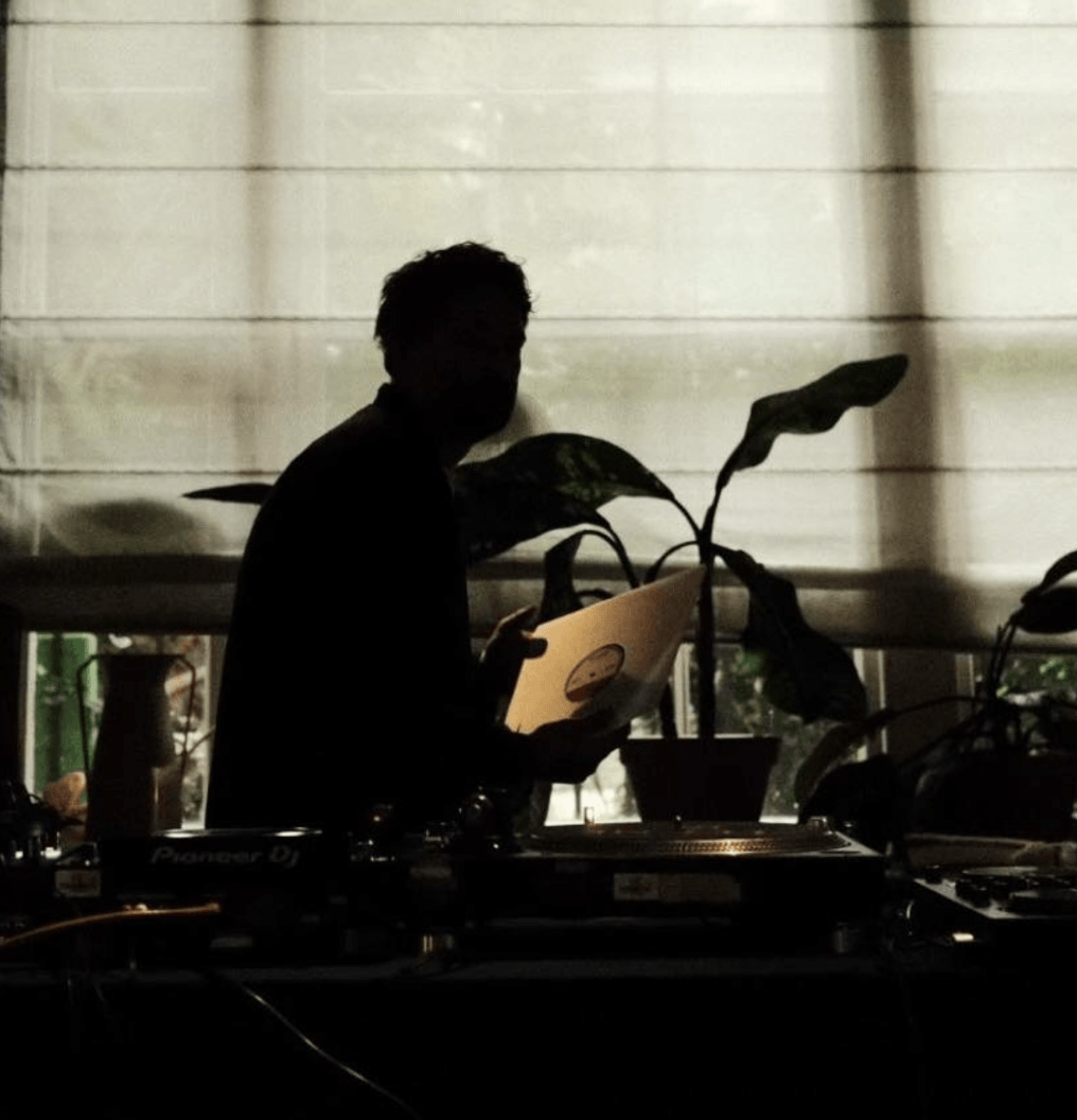 Silhouette of a person holding a record album behind a DJ table with plants in the background.