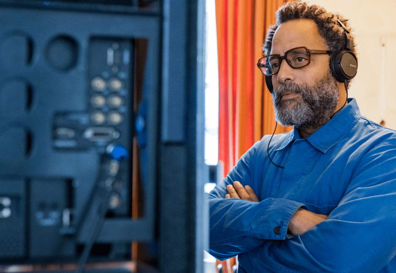 A man with glasses and a gray beard wearing headphones and a blue shirt, intently watching a computer monitor.