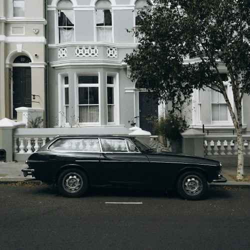 A vintage black car parked on a city street in front of a white Victorian-style house with decorative trim and railings.