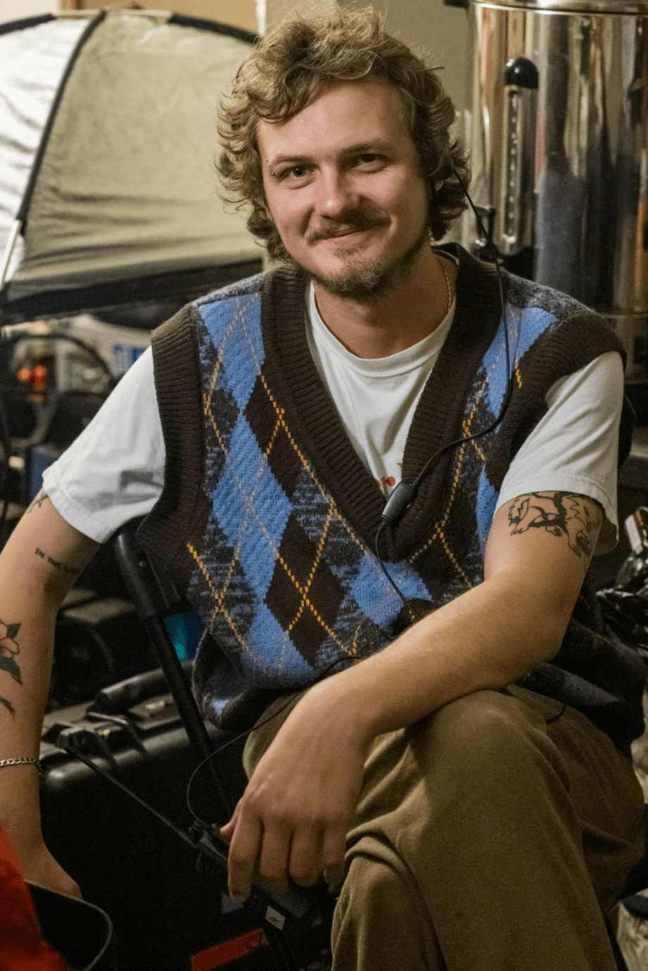 A man with curly hair, a mustache, and tattoos on his arms, sitting and smiling, wearing a white T-shirt and a blue and brown plaid vest, with a camera and equipment around him.