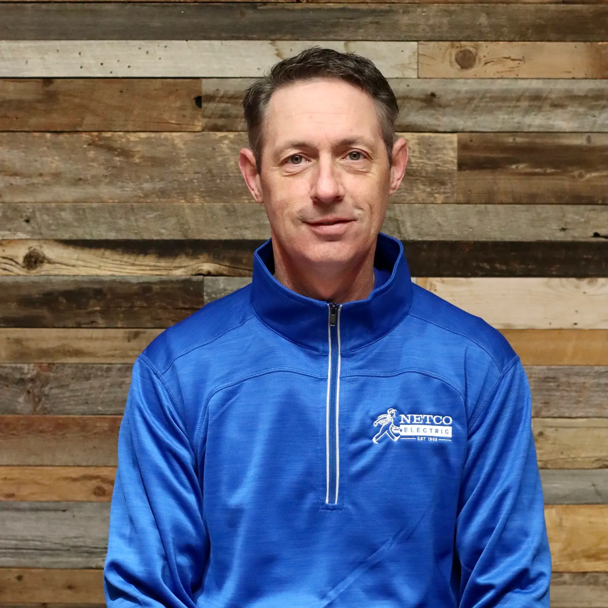 A man in a blue NETCO Electric jacket standing in front of a wooden wall.
