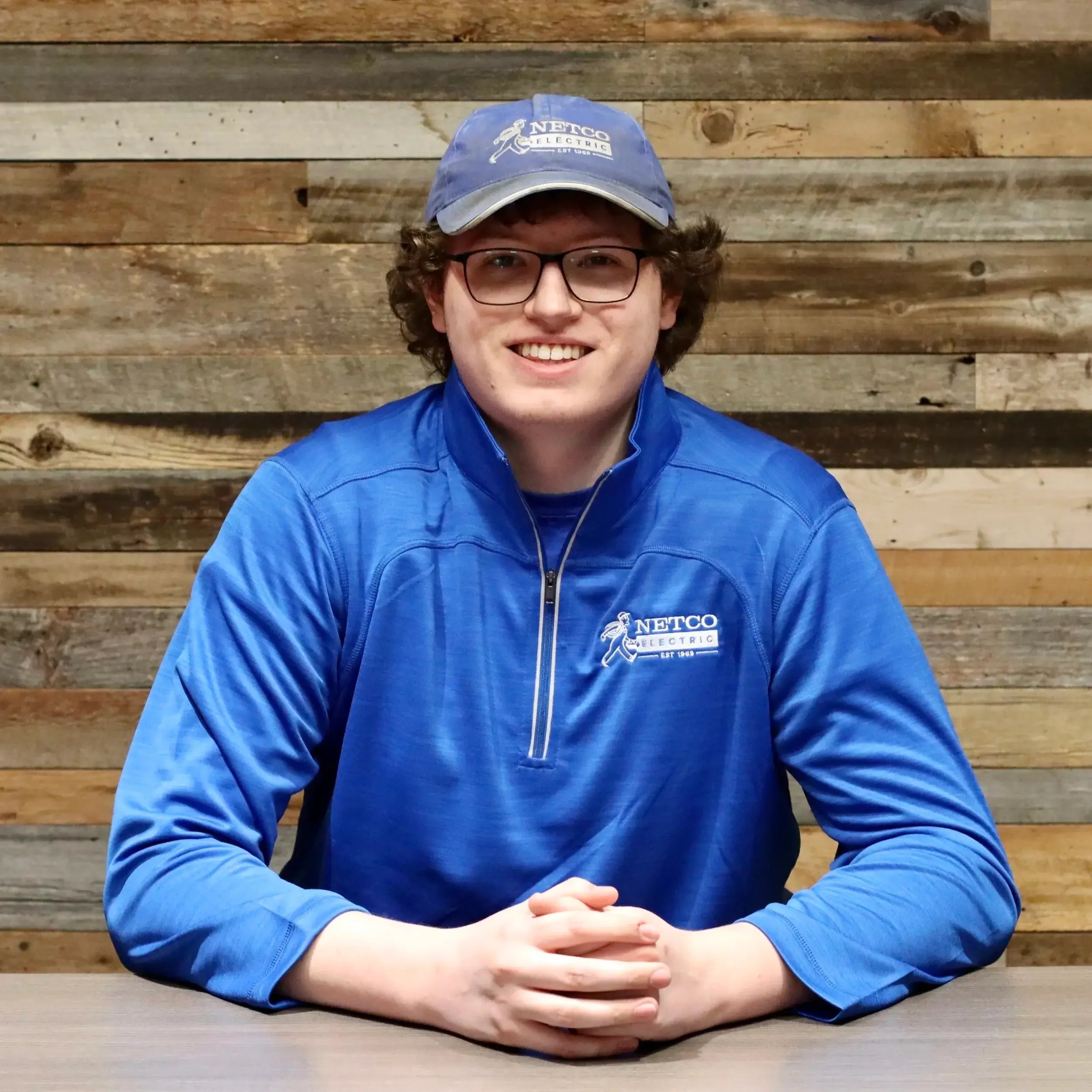A young man with curly hair, glasses, and a wide smile, wearing a blue NETCO Electric cap and a matching blue pullover, sitting at a table in front of a wooden plank wall.