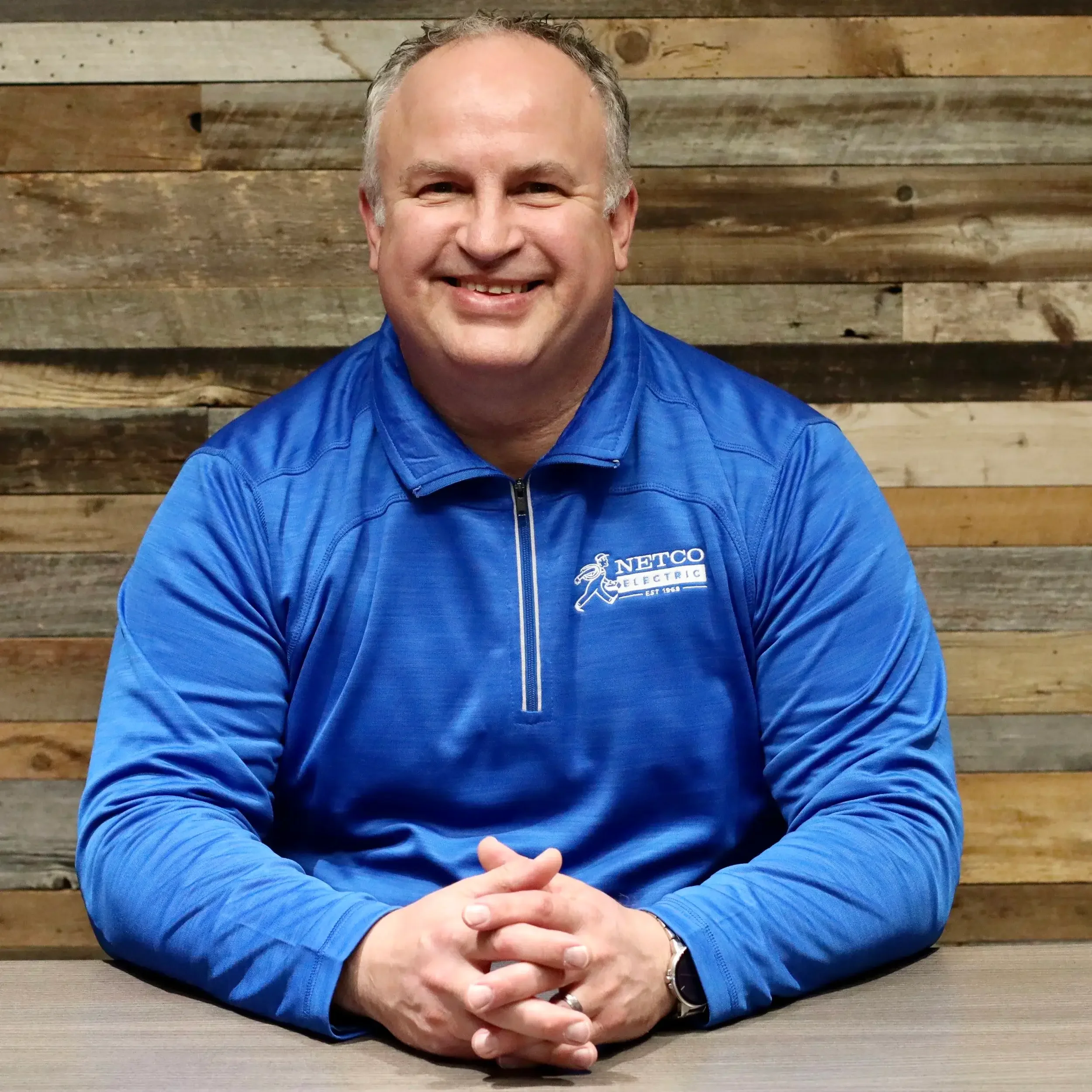 A smiling man with short gray hair wearing a blue jacket with a 'NETCO Electrical' logo, sitting at a table in front of a wooden wall.