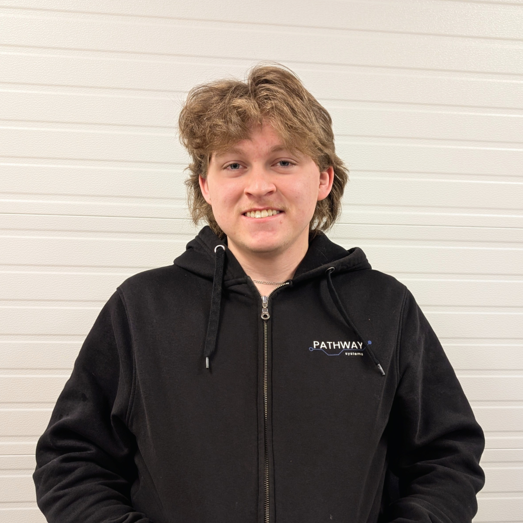 A young man with brown hair and braces, wearing a blue jacket with a logo that says 'NETCO Electric,' standing in front of a wooden wall.