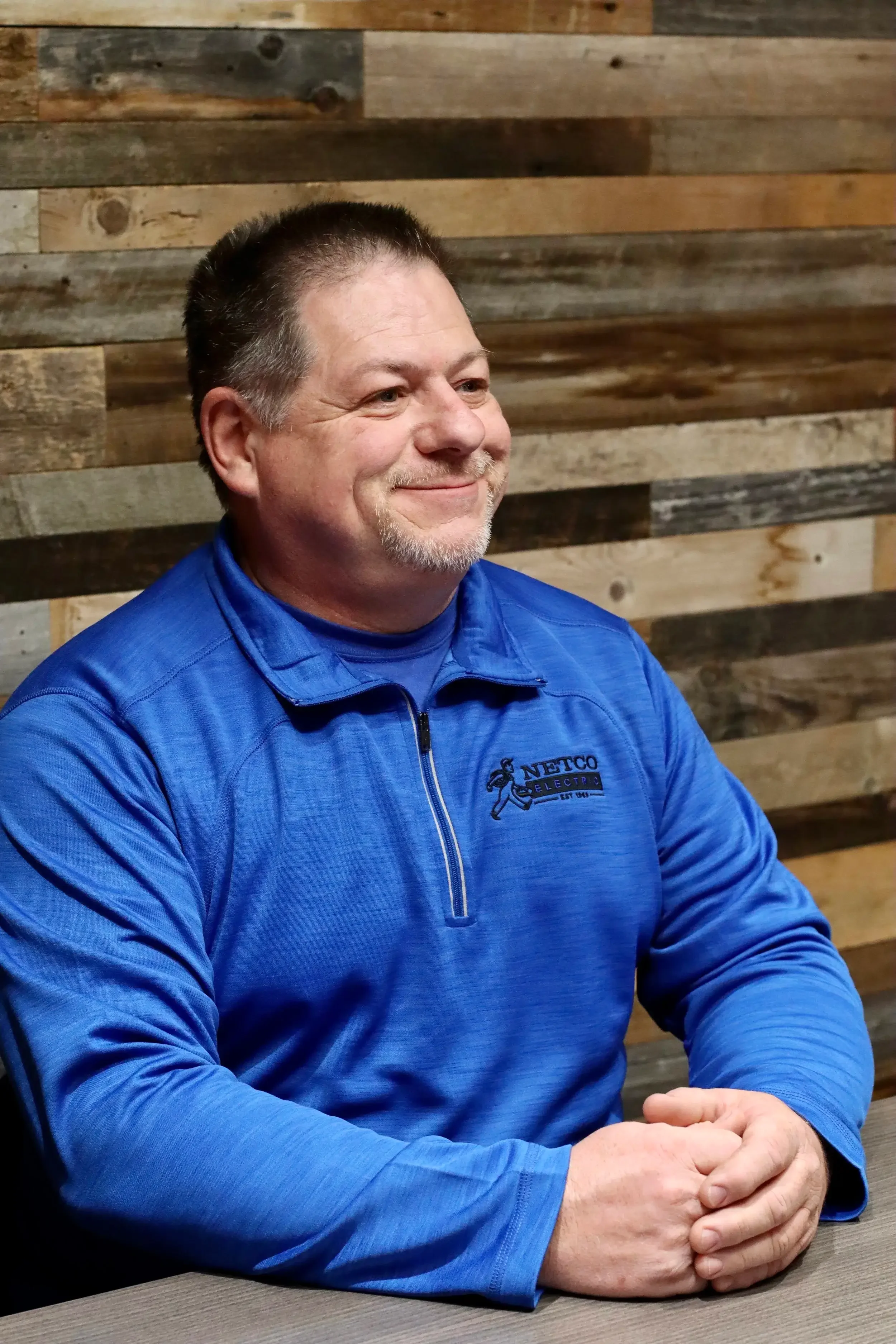 A smiling man wearing a blue sweatshirt with a logo, sitting at a table with a wooden plank wall in the background.
