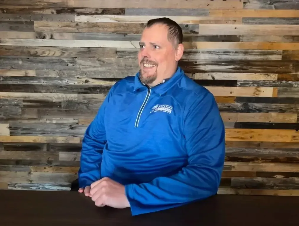 A man with short dark hair and a beard, wearing a blue jacket, smiling and sitting at a table with a rustic wooden wall in the background.