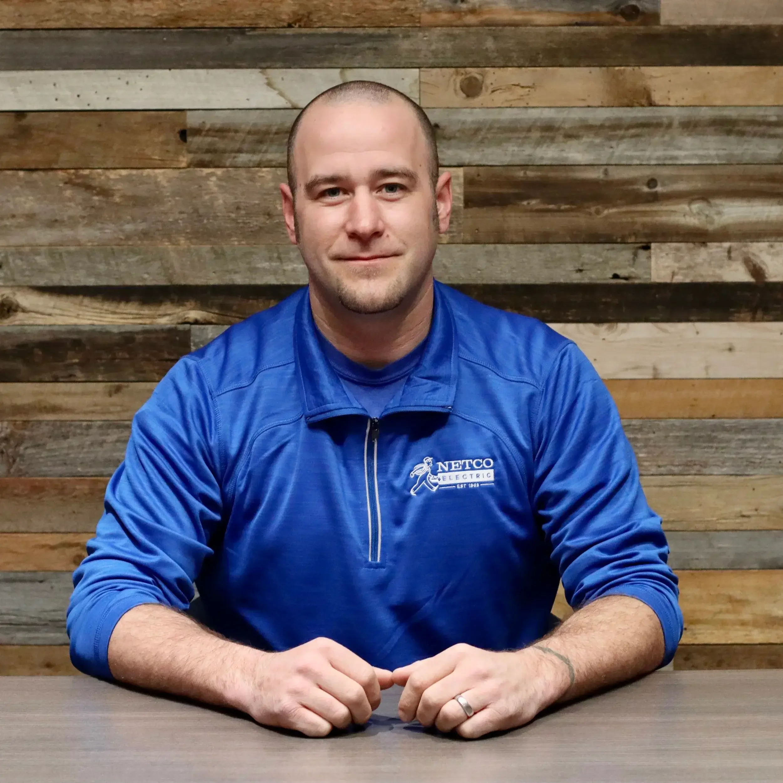 A man with a shaved head wearing a blue shirt with a logo reading 'NETCO Electric' sitting at a table with a wood-paneled wall background.
