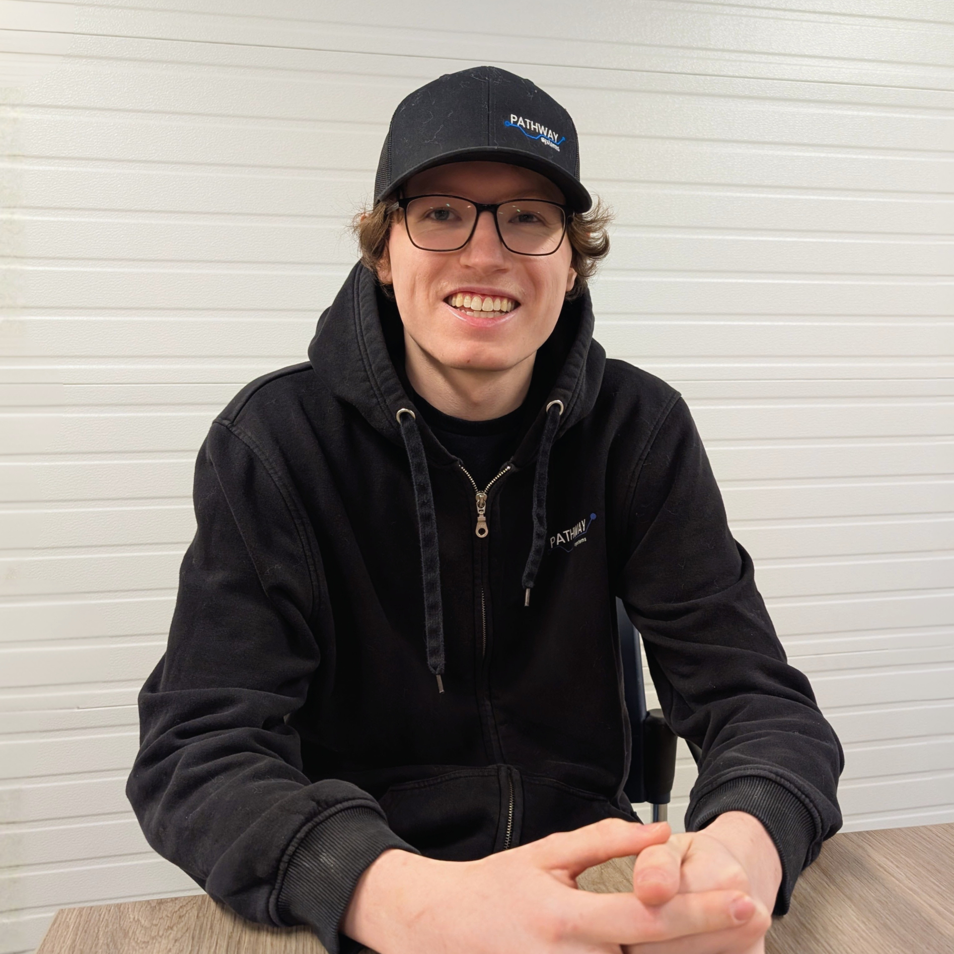 A young man with curly hair, glasses, and a wide smile, wearing a blue NETCO Electric cap and a matching blue pullover, sitting at a table in front of a wooden plank wall.