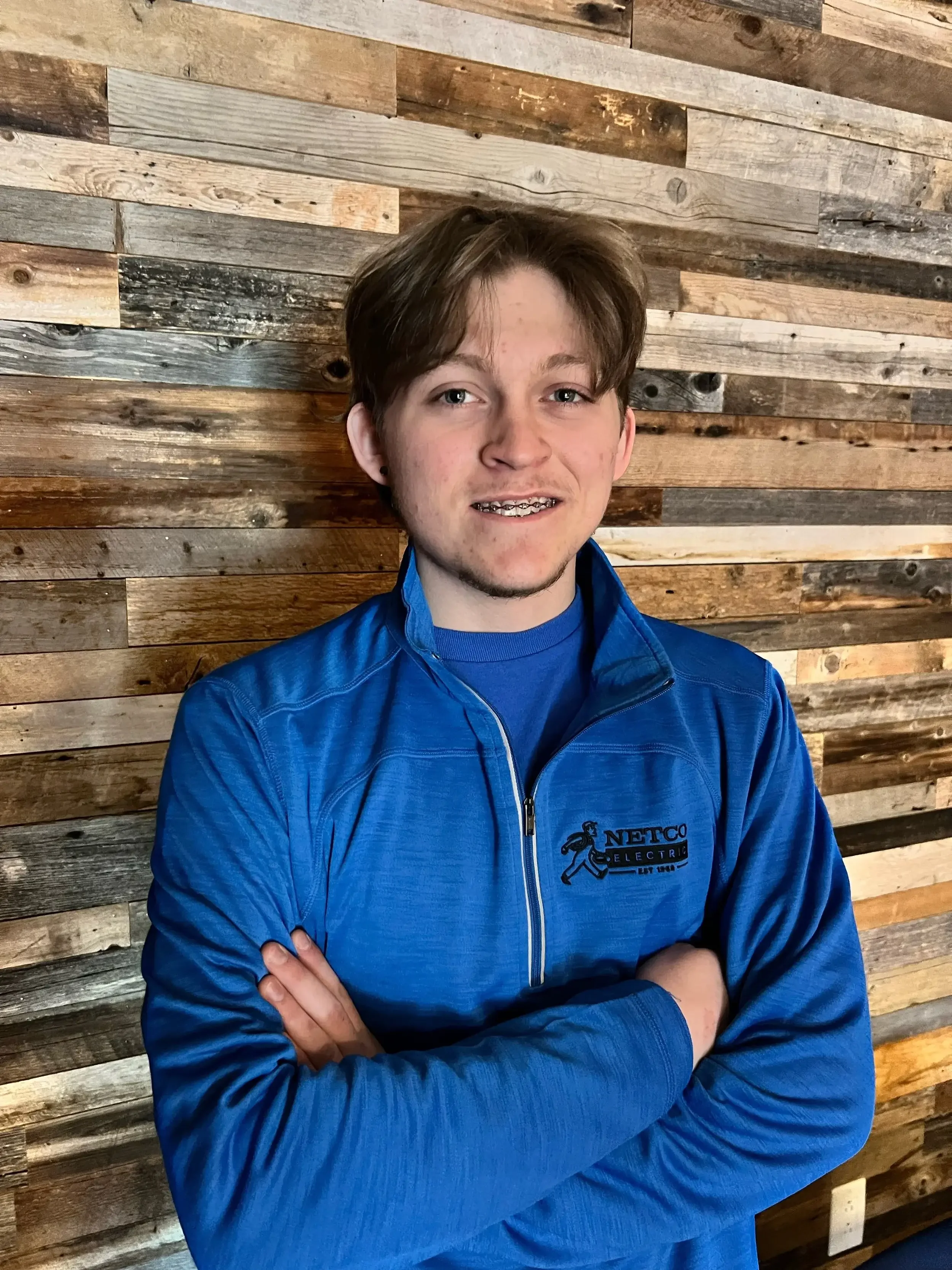 A young man with brown hair and braces, wearing a blue jacket with a logo that says 'NETCO Electric,' standing in front of a wooden wall.