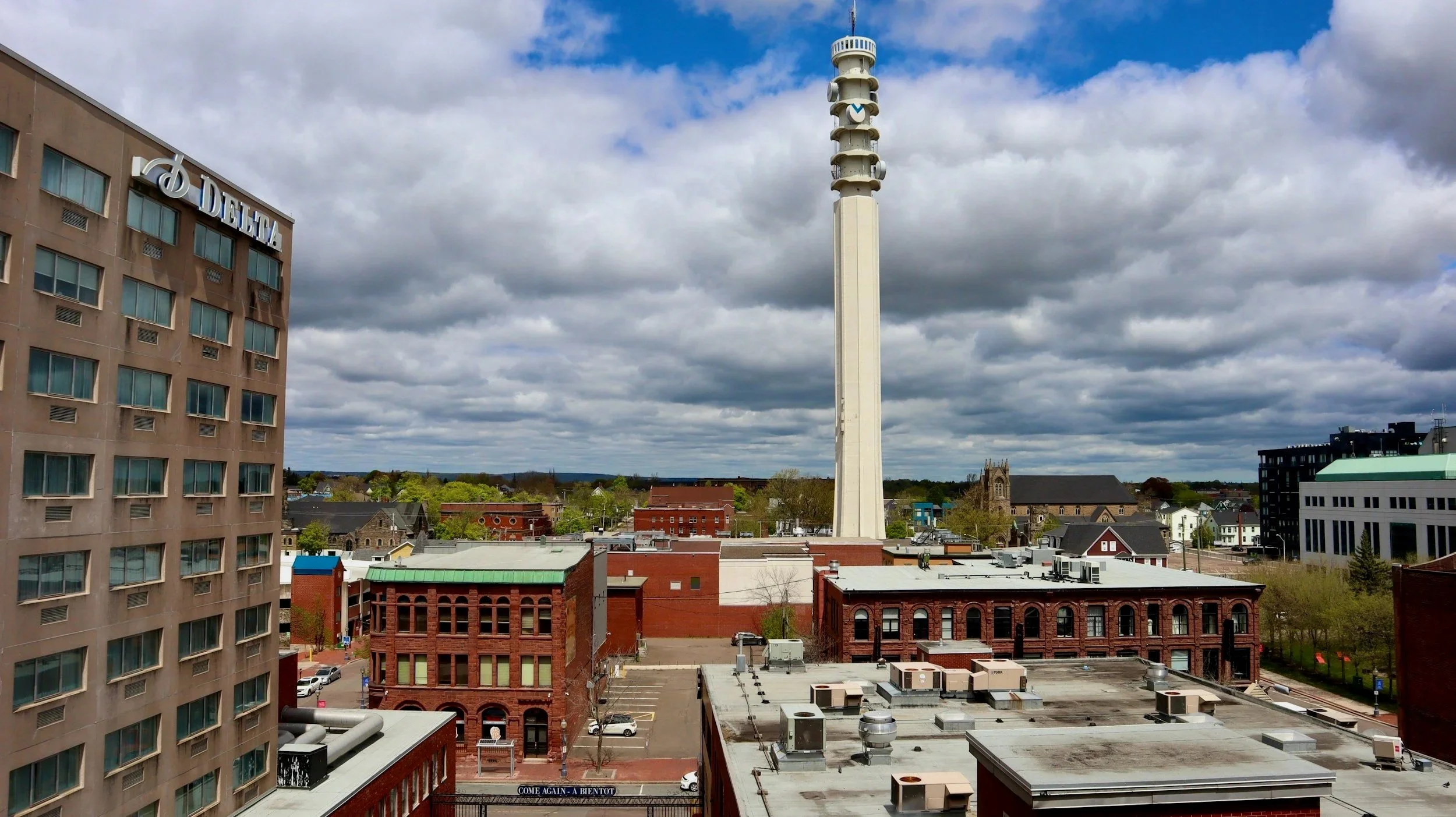 Cityscape with a tall white observation tower under a cloudy sky, surrounded by multi-story buildings and trees.