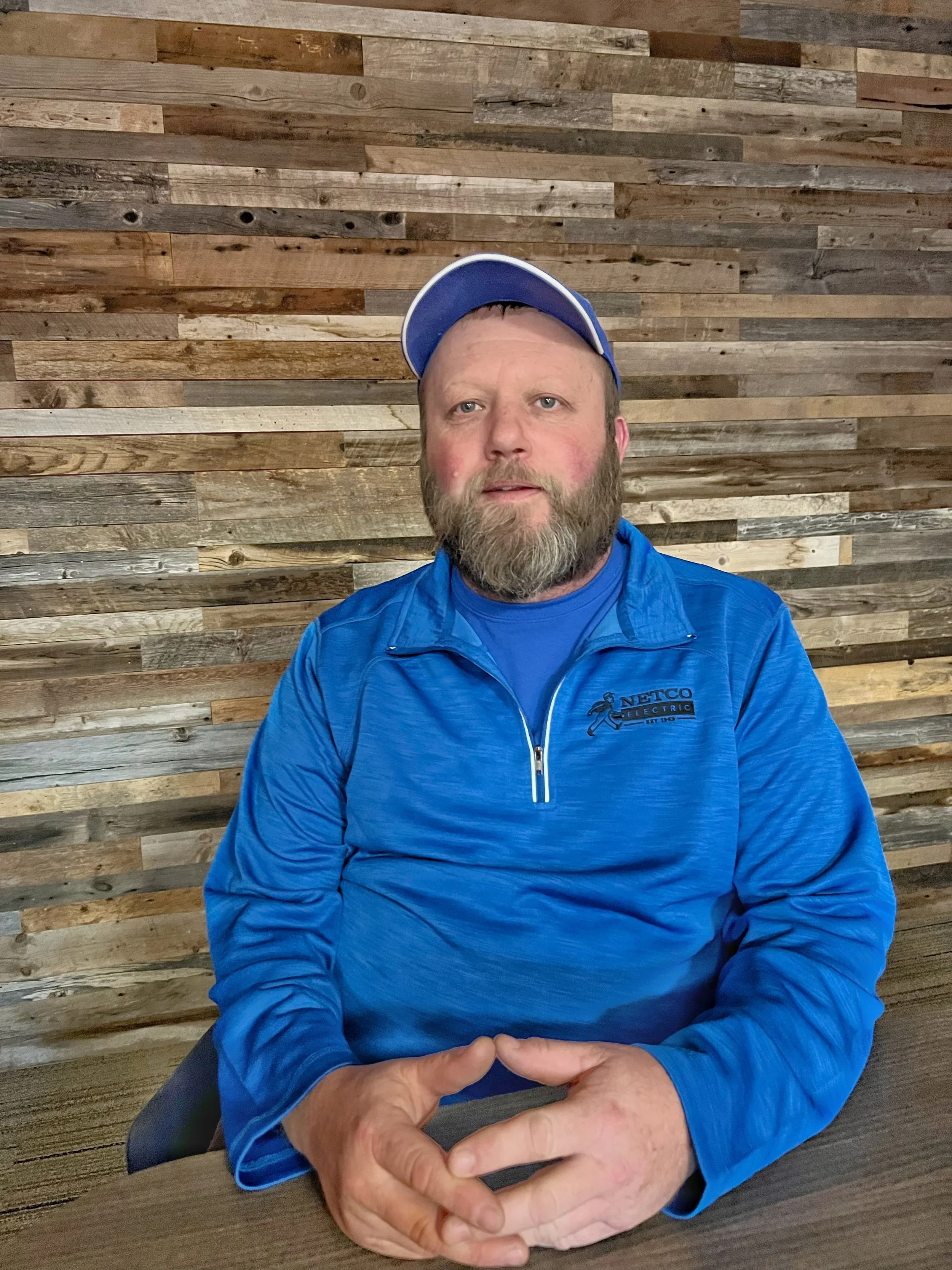 A man with a bald head and a beard, smiling, sitting at a table in front of a wooden wall background, wearing a blue jacket with a 'NETCO Electric' logo.