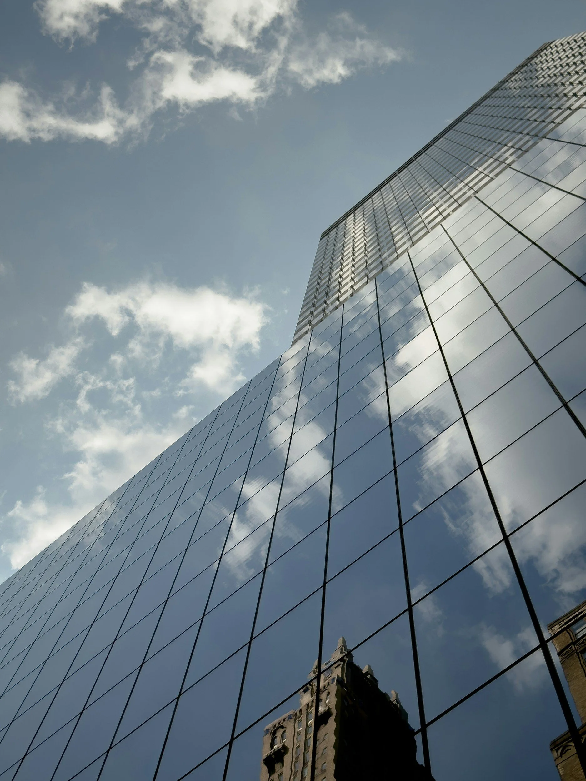 Looking up at a modern glass skyscraper reflecting the sky and clouds, with a reflection of an older building on the glass surface.
