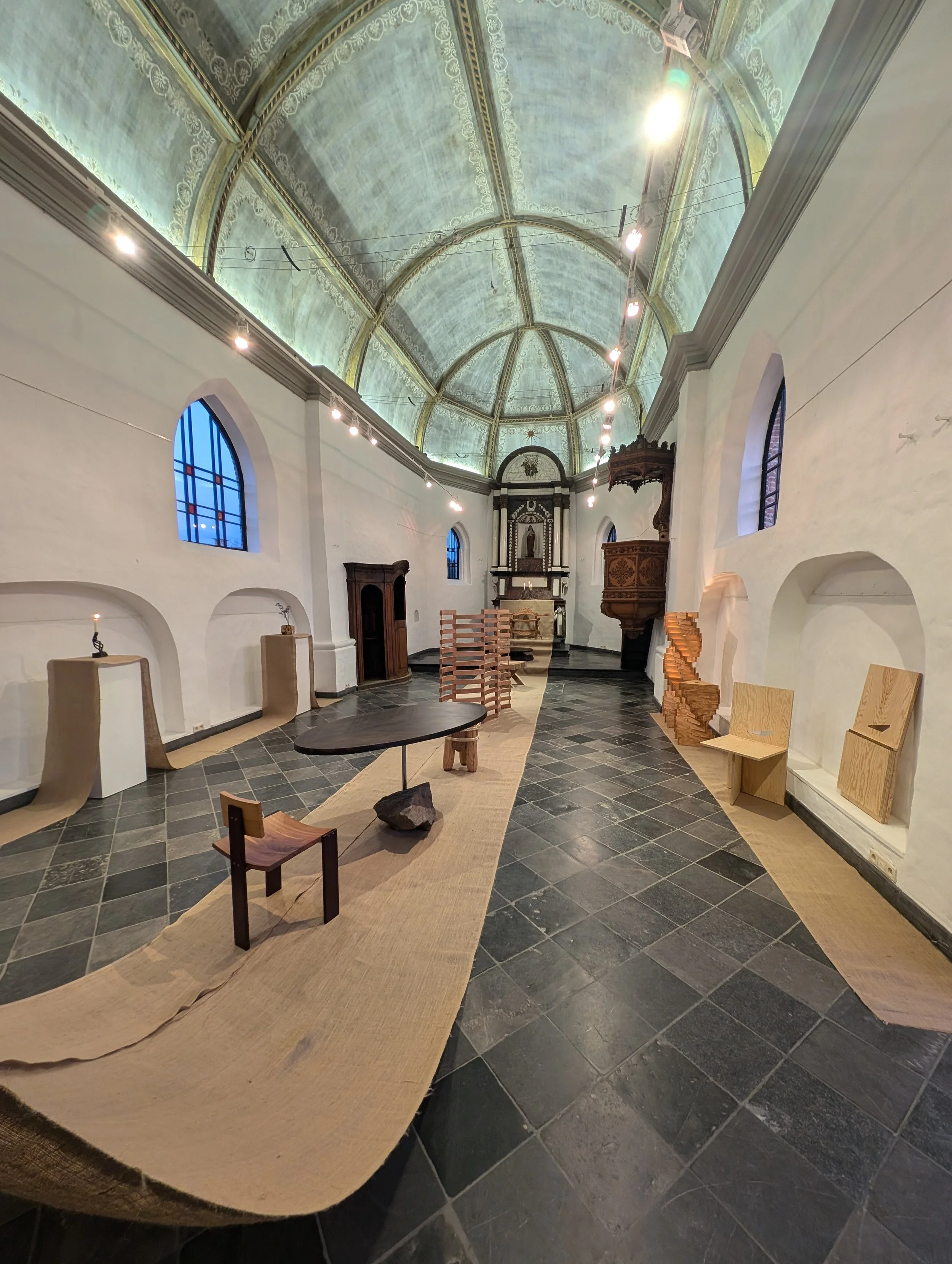Interior of a historical church with rustic furniture and modern art installations, high arched ceiling, stained glass windows, and a tiled floor.