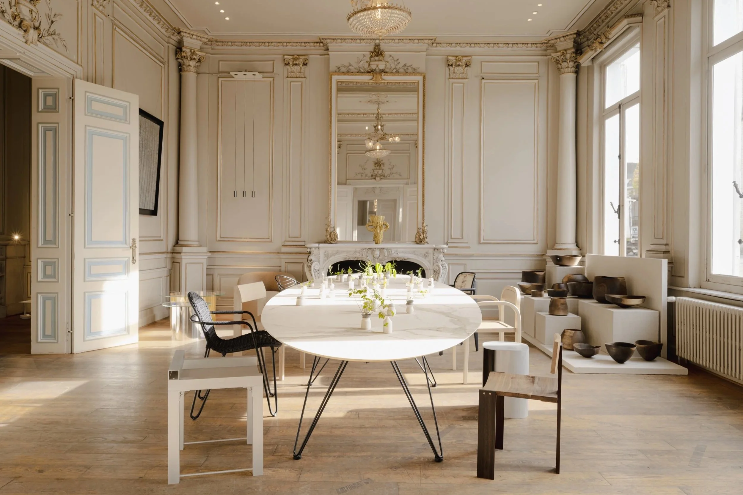 An elegant, spacious room with high ceilings, large windows, decorative molding, and a large mirror above a white ornate fireplace. The room contains a long oval white table with small potted green plants and chairs of different styles around it, as well as displayed wooden bowls and sculptures near the windows.