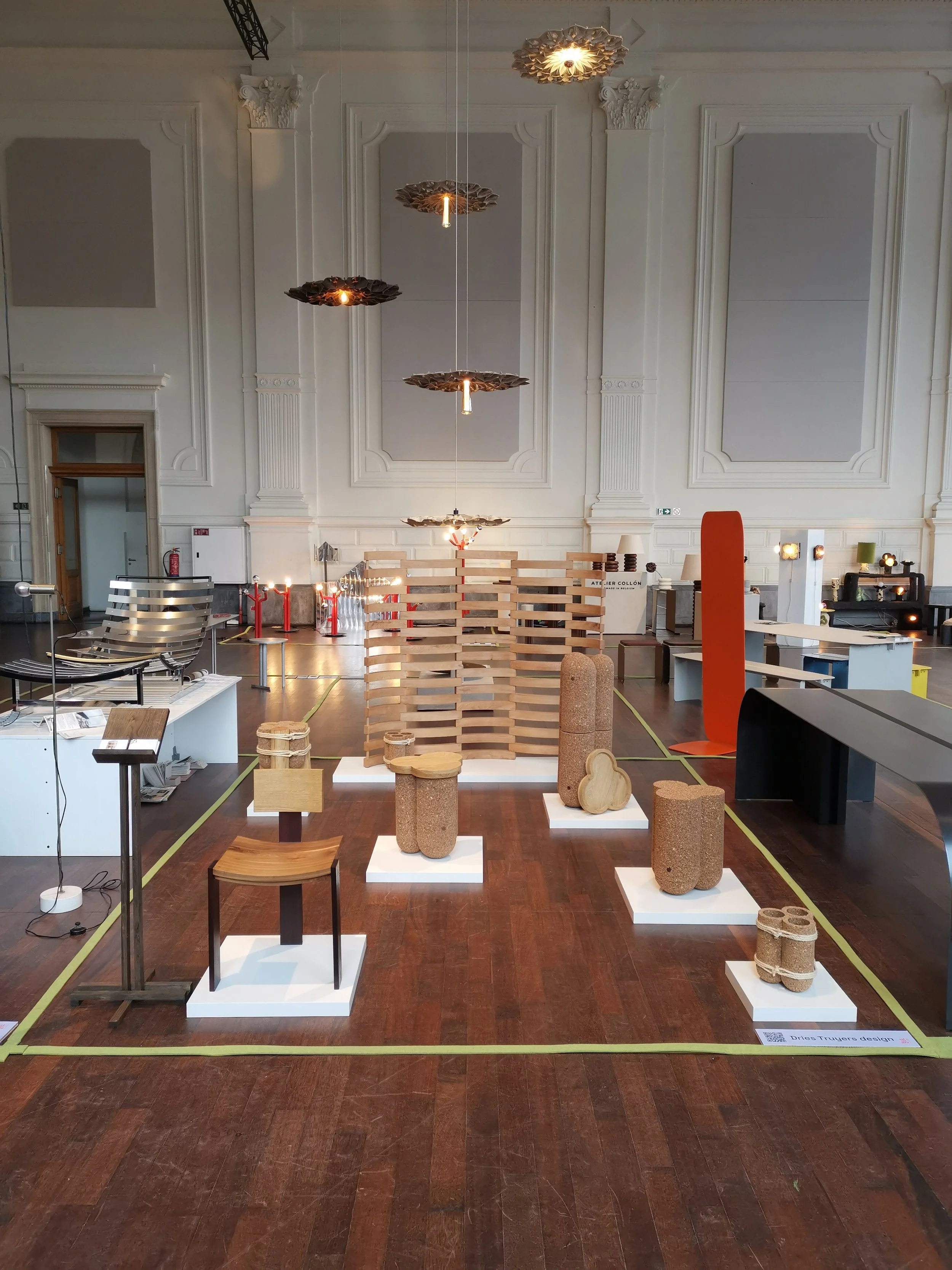 Art display in a gallery with various wooden and cork sculptures and furniture pieces on white pedestals, a wooden partition, and modern lighting fixtures in a spacious, high-ceiling room.