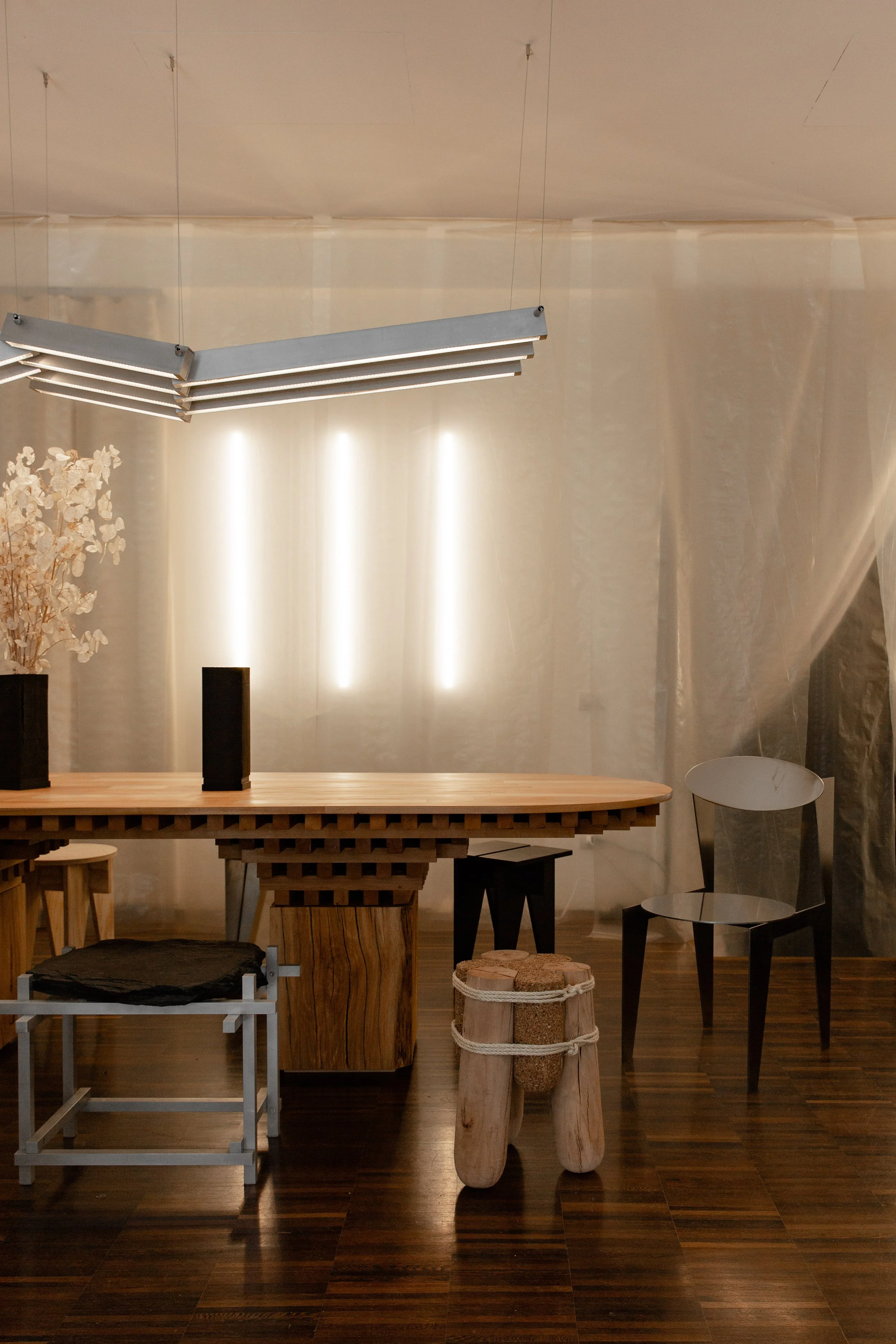 A modern interior room with a wooden table, black and wooden chairs, decorative vases, and unique furniture pieces, illuminated by overhead lighting.
