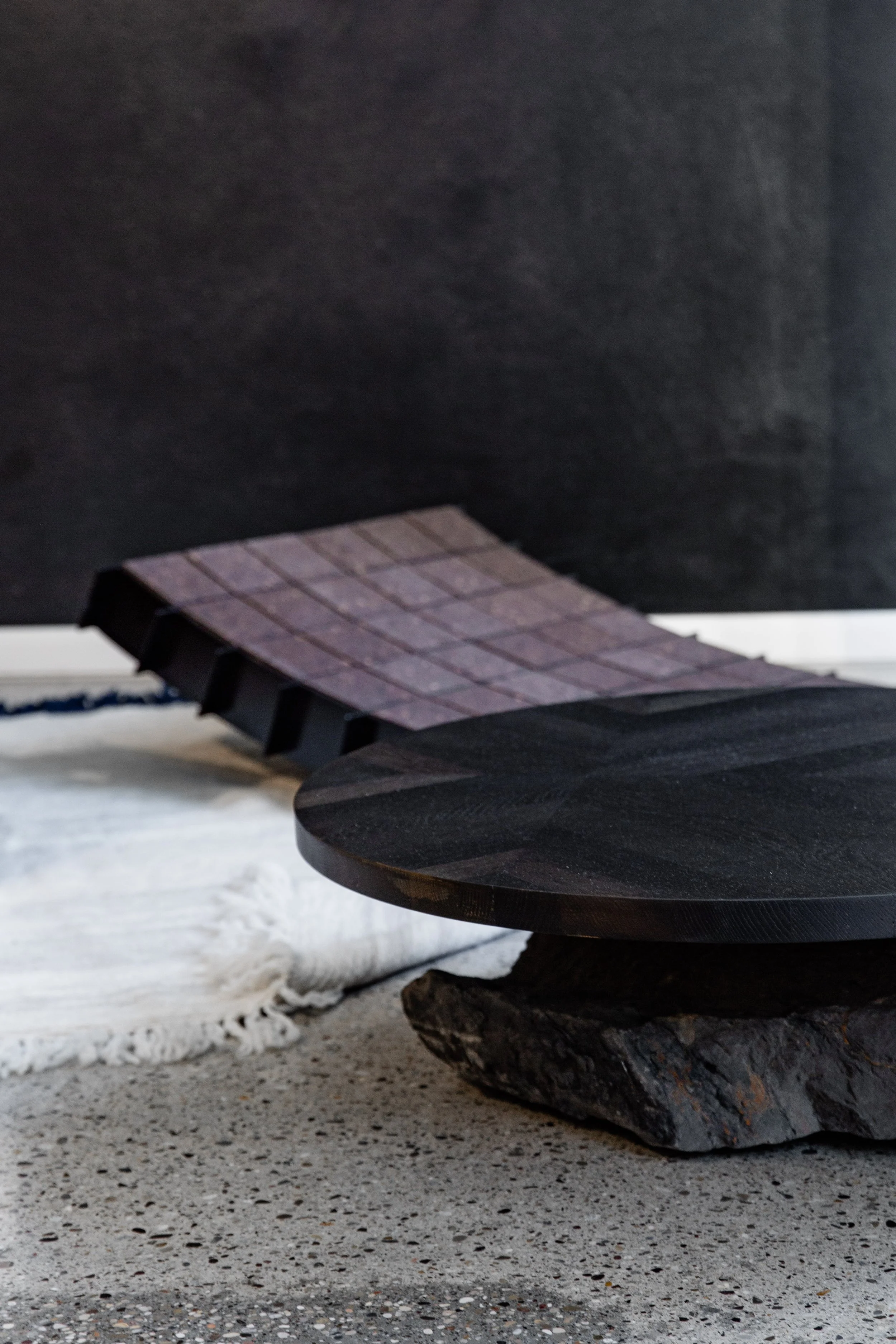 A modern, minimalist coffee table made of a large, flat, dark stone slab resting on a rough, textured stone base, with a black wall in the background, a white rug underneath, and a black bookshelf or wall behind.